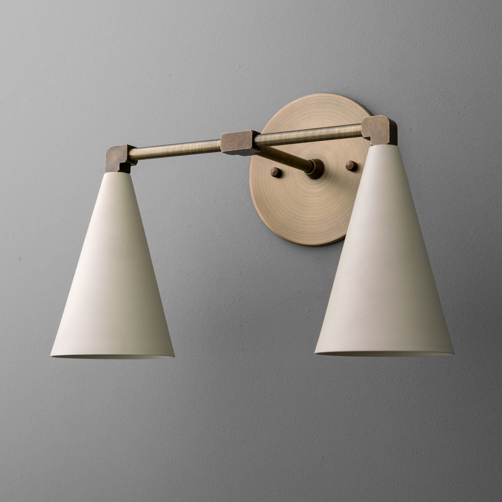 Angled view of the two-light vanity light in Antique Brass with Cream cone shades on a straight arm and round wall plate. - VANITY LIGHT MODEL NO. 1184
