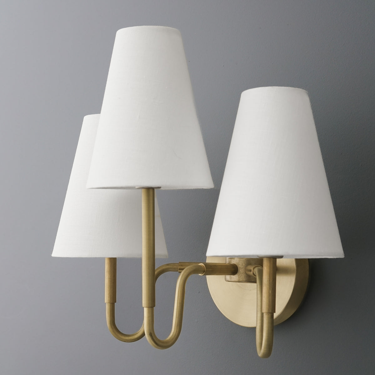 Three-bulb wall sconce with brushed brass arms and round backplate, featuring white cone-shaped fabric shades. - SCONCE MODEL NO. 4940