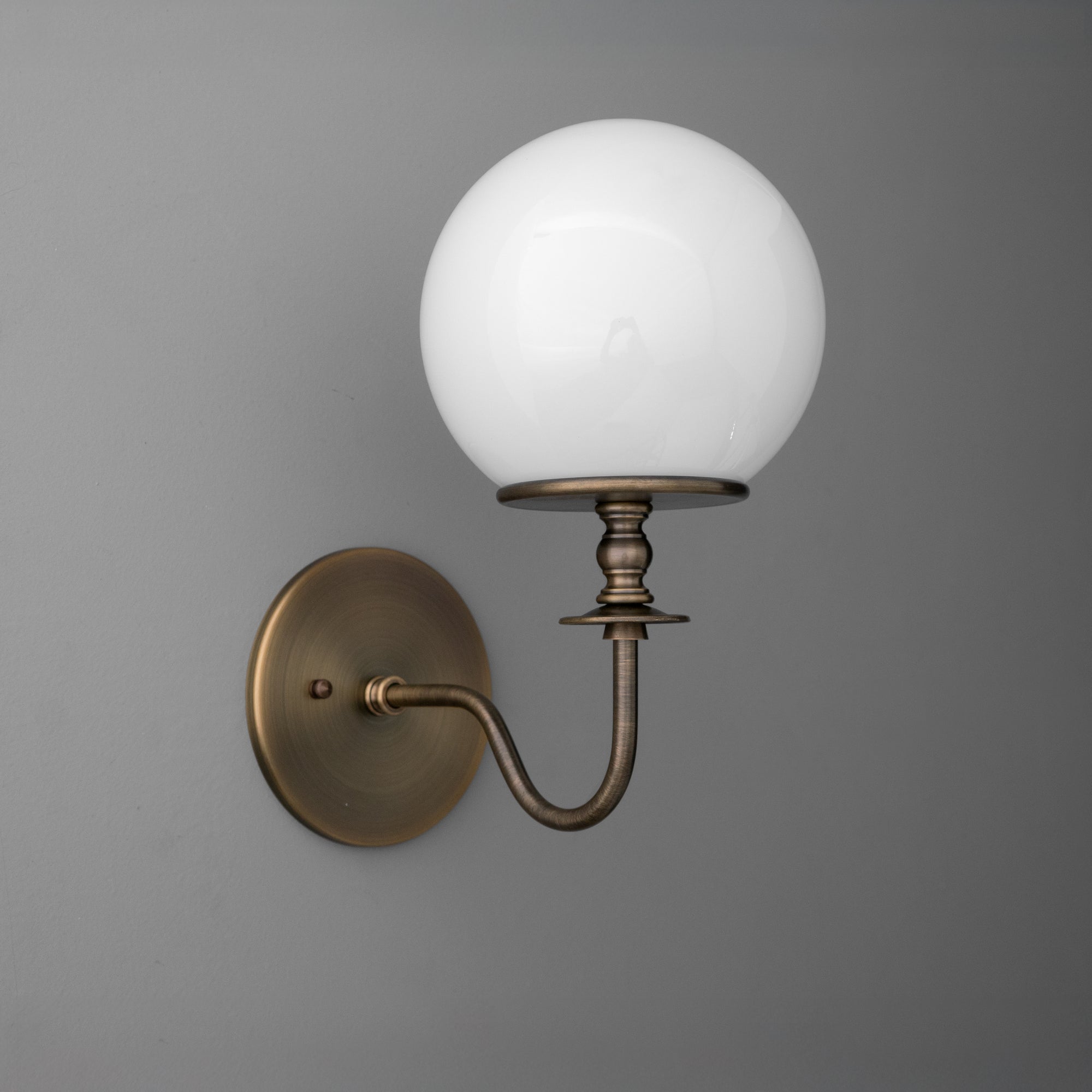 Antique brass wall sconce with curved arm and white glass globe shade on gray background. - SCONCE MODEL NO. 7857