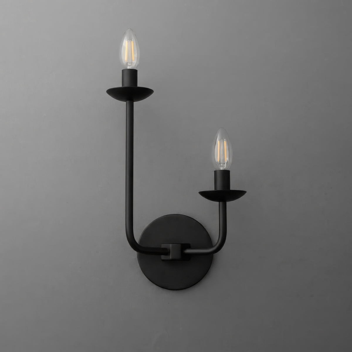 Two-light sconce in Black with the long arm on the left and the short arm on the right, each with a candle cup and exposed bulb, on a round wall plate. - SCONCE MODEL NO. 9725