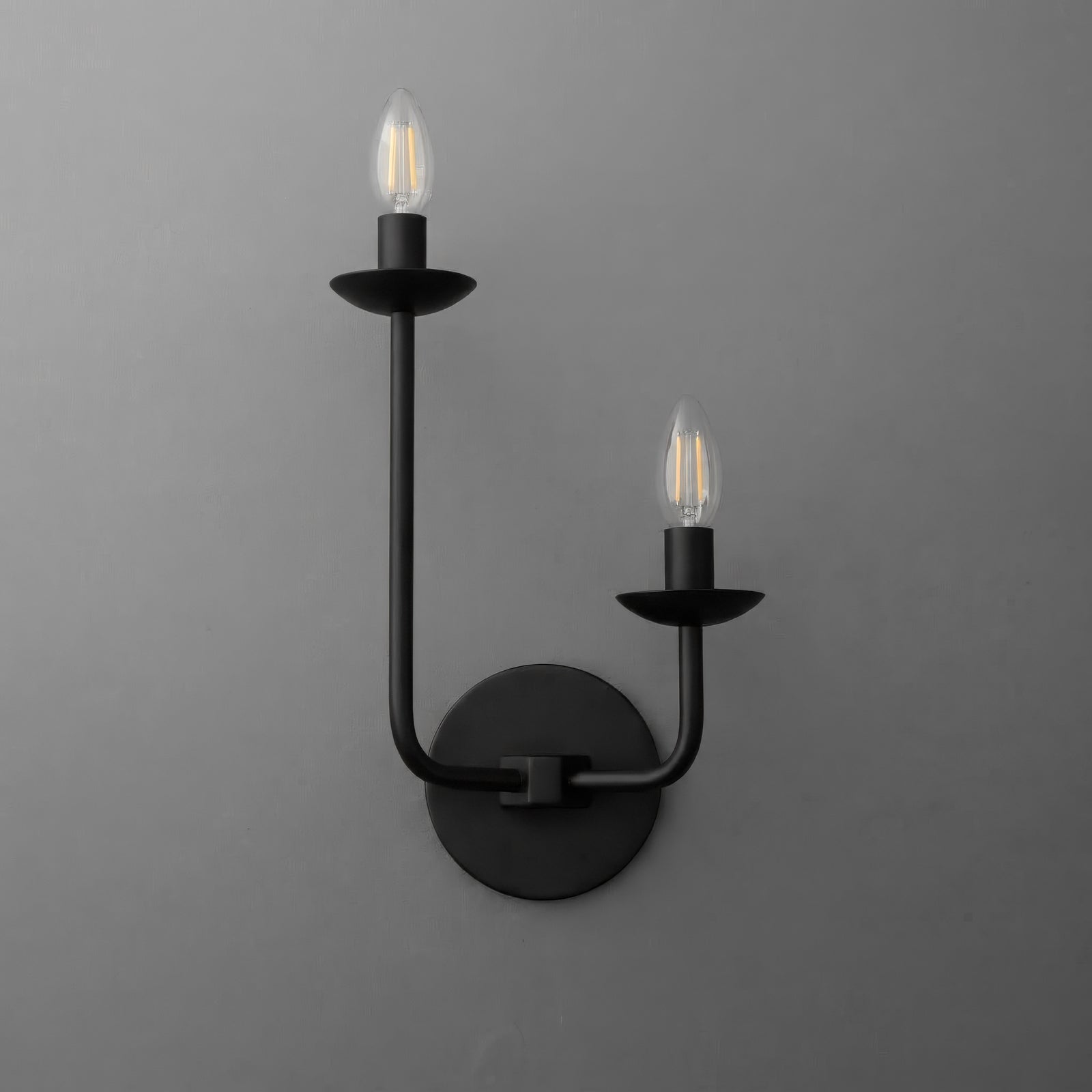 Two-light sconce in Black with the long arm on the left and the short arm on the right, each with a candle cup and exposed bulb, on a round wall plate. - SCONCE MODEL NO. 9725