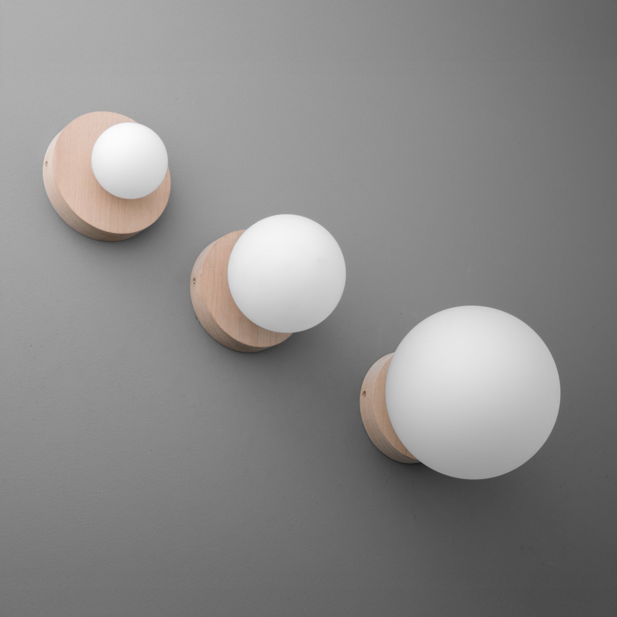 Three modern wall sconces with beechwood bases and frosted white glass globes in small, medium, and large sizes on a gray. - SCONCE MODEL NO. 4943