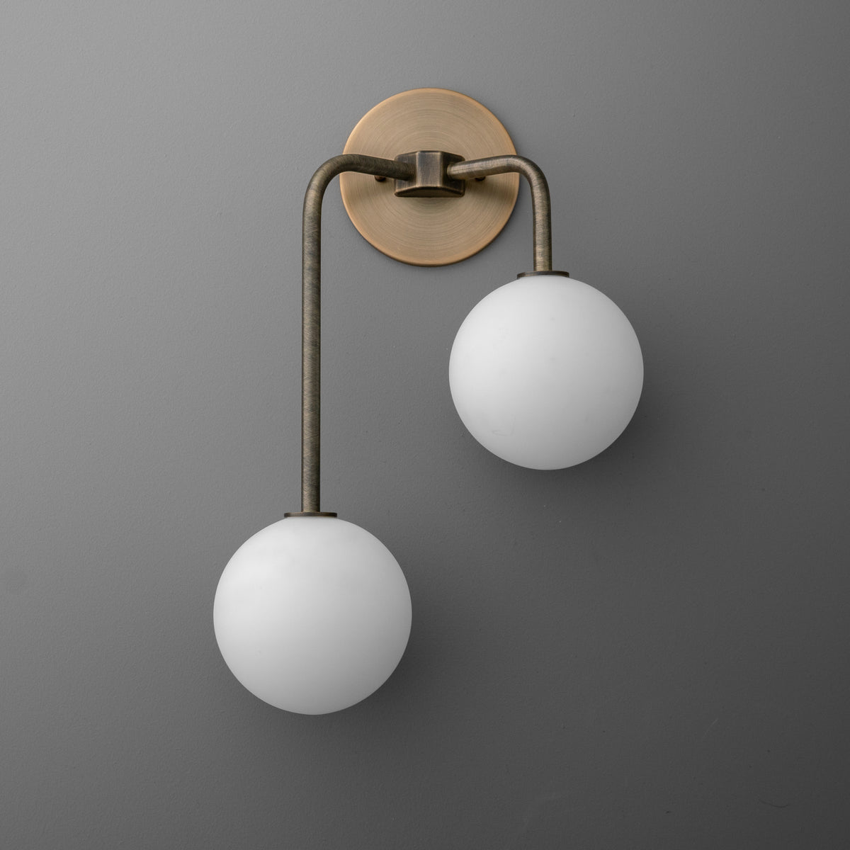 Two-light sconce in Antique Brass with opal glass globes, the long arm on the left holding the lower globe, on a round wall plate. - SCONCE MODEL NO. 3996