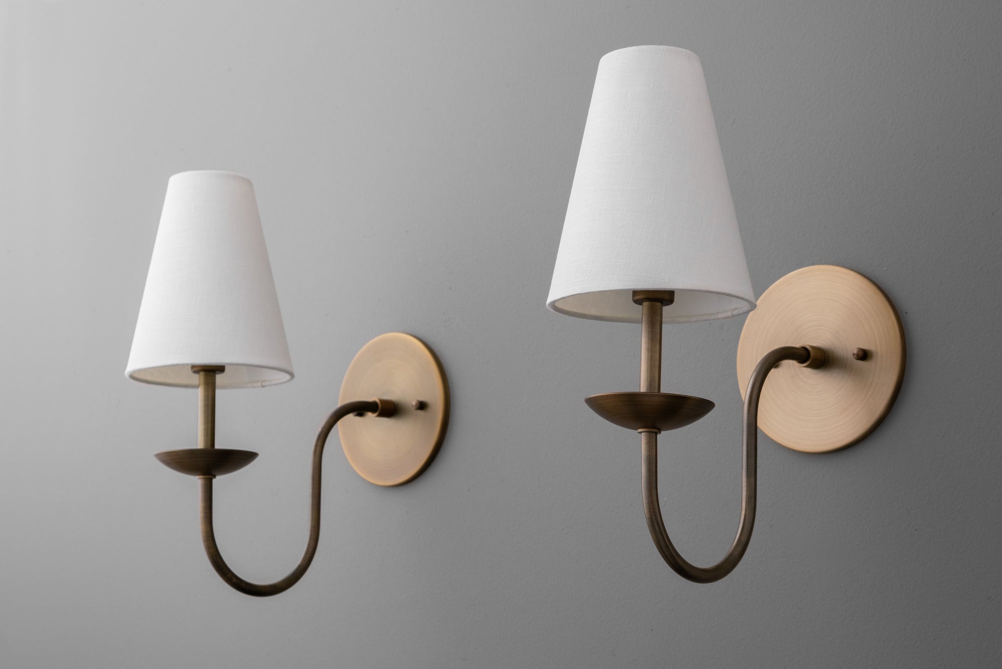 Pair of antique brass sconces with white tapered fabric shades and curved arms mounted on round backplates. - SCONCE MODEL NO. 9867