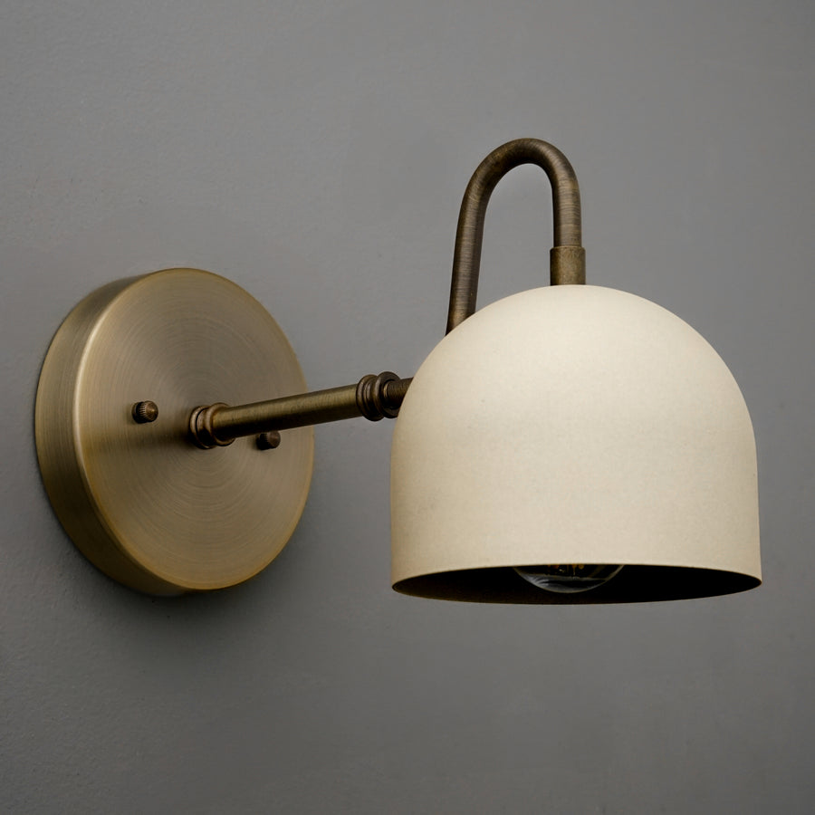 Antique brass wall sconce with cream dome-shaped metal shade and curved arm mount on gray wall. - SCONCE MODEL NO. 3628