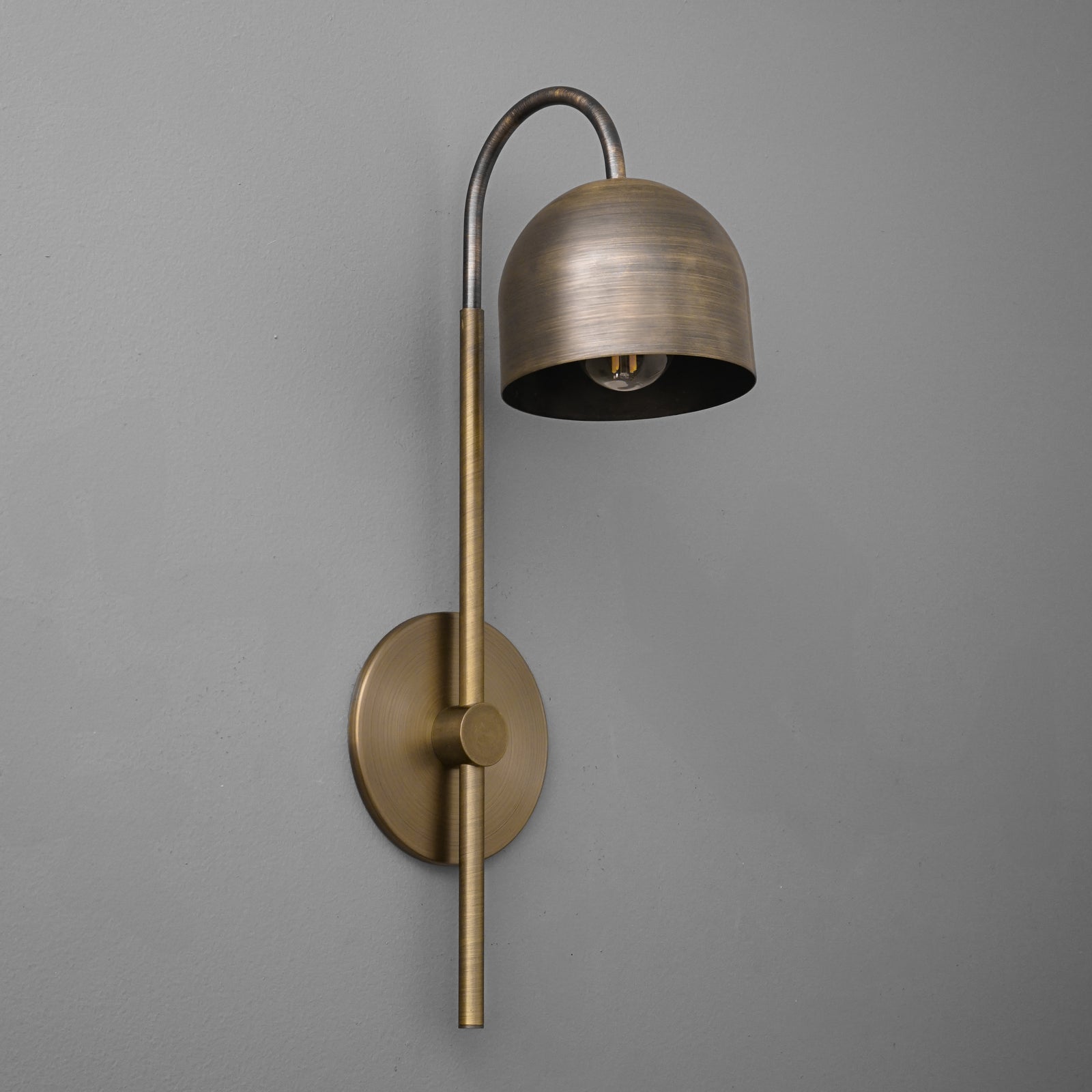 Antique brass wall sconce with curved arm and dome-shaped shade against gray wall. - Sconce Model No. 7340