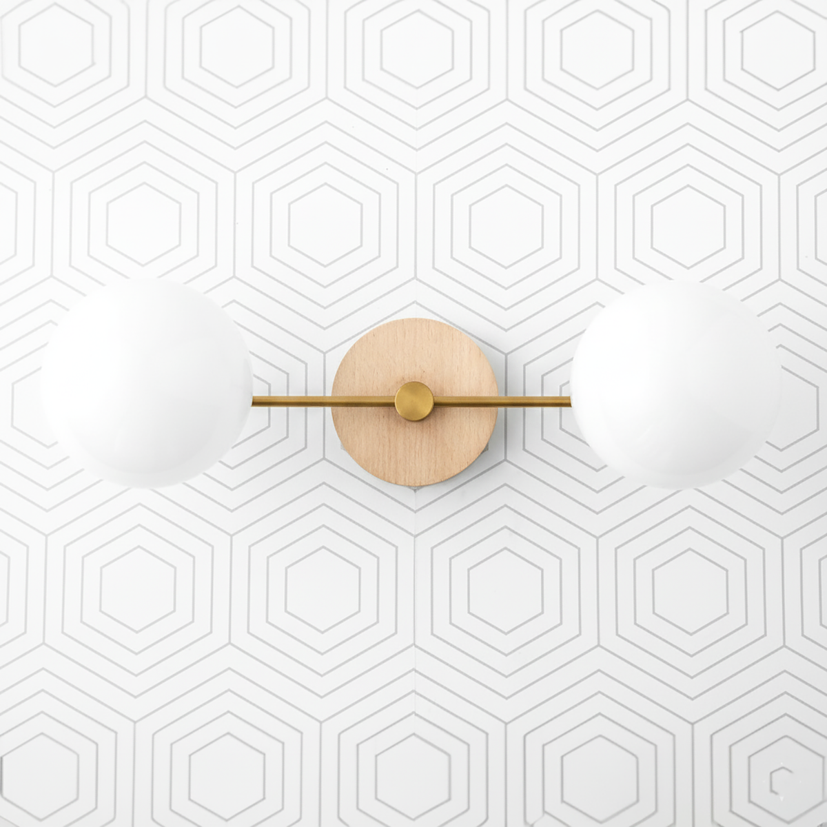 Two-globe vanity light with raw brass horizontal bar and natural wood round wall mount against hexagon patterned white wall. - VANITY LIGHT MODEL NO. 9474