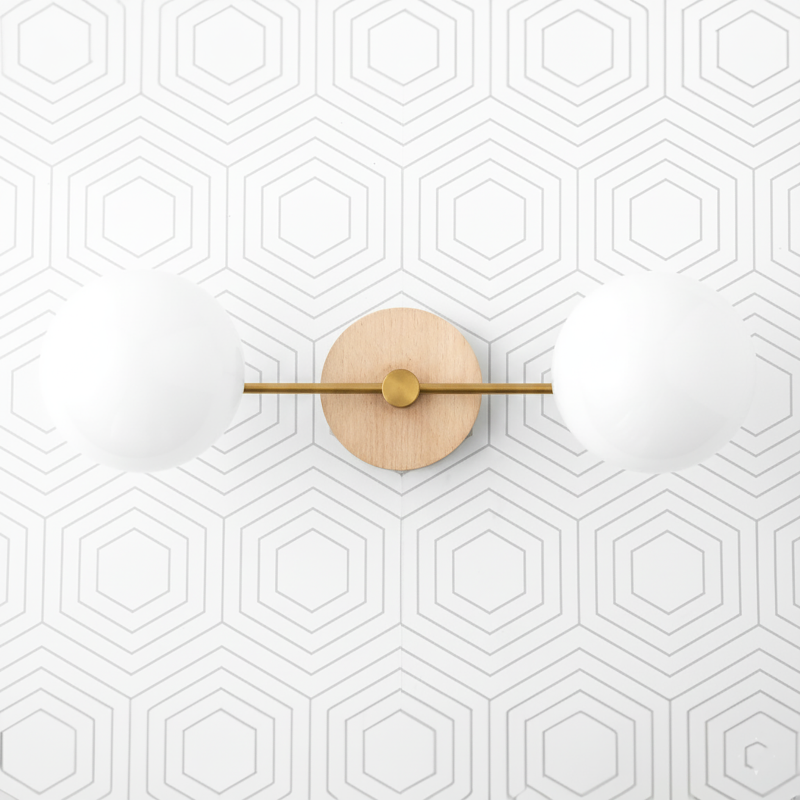 Two-globe vanity light with raw brass horizontal bar and natural wood round wall mount against hexagon patterned white wall. - VANITY LIGHT MODEL NO. 9474