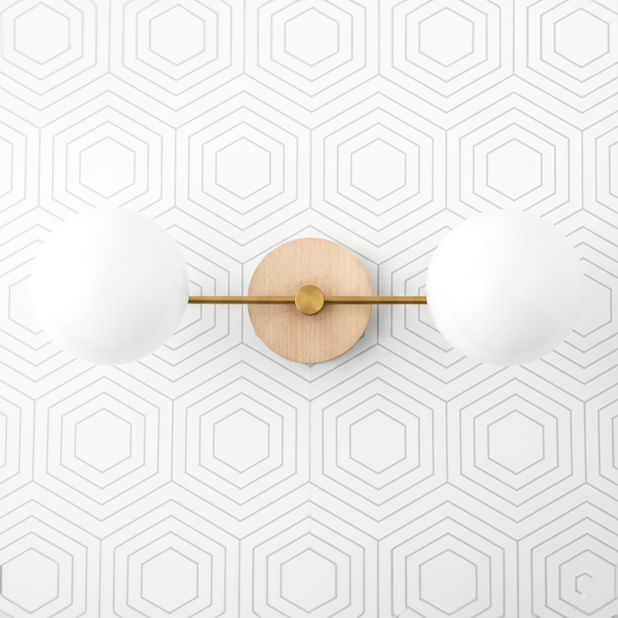 Two-globe vanity light with raw brass horizontal bar and natural wood round wall mount against hexagon patterned white wall. - VANITY LIGHT MODEL NO. 9474