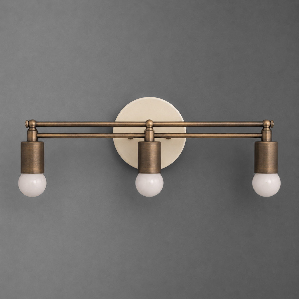 Three-light vanity light in Antique Brass with cylinder sockets and exposed globe bulbs hanging from a double bar on a Cream round wall plate. - VANITY LIGHT MODEL NO. 8256