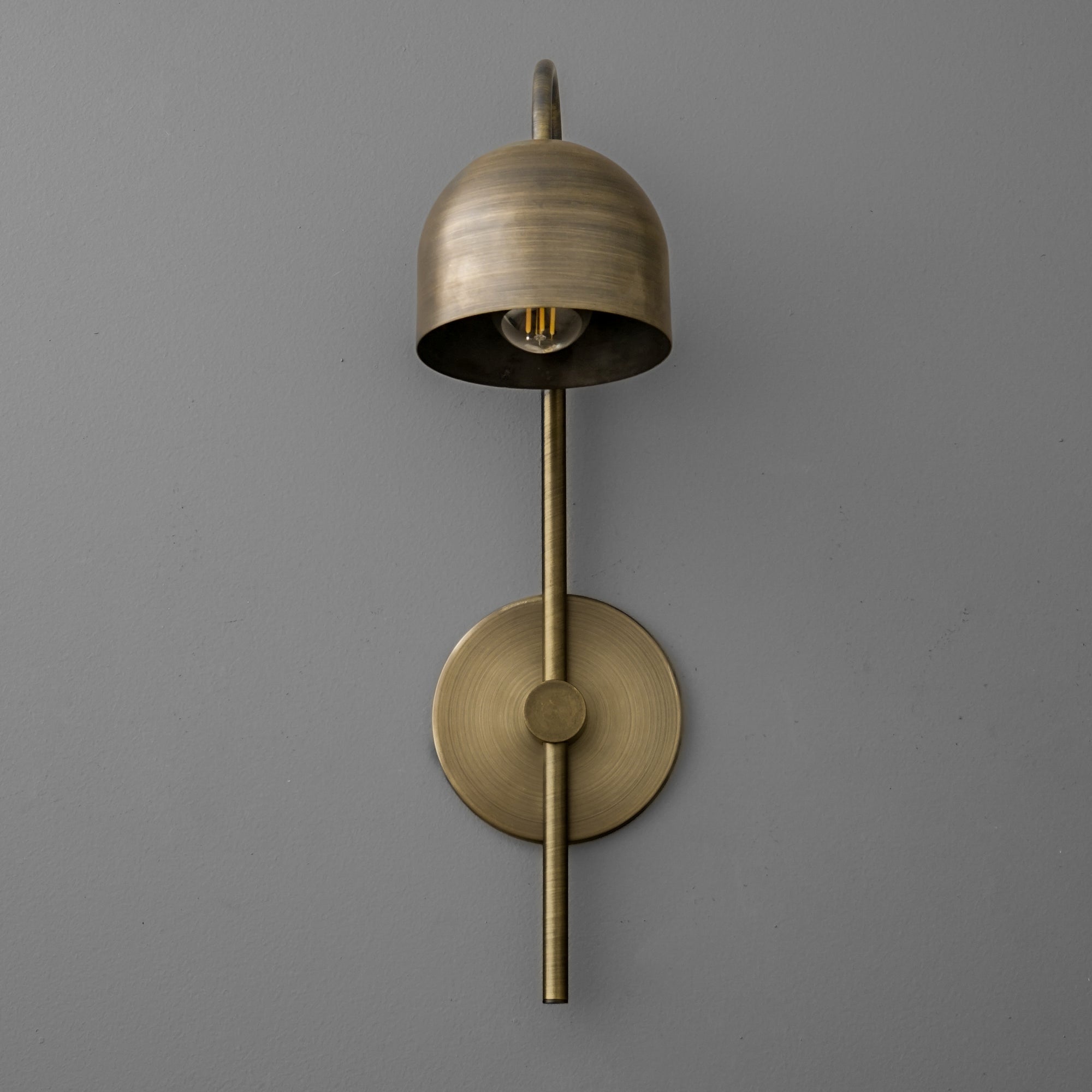 Antique brass sconce with bell-shaped shade and vertical rod mounting on gray wall. - Sconce Model No. 7340