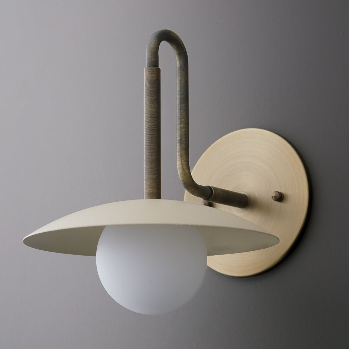 Matte beige metal wall sconce with curved arm and wide flat shade over frosted glass globe bulb. - SCONCE MODEL NO. 1017