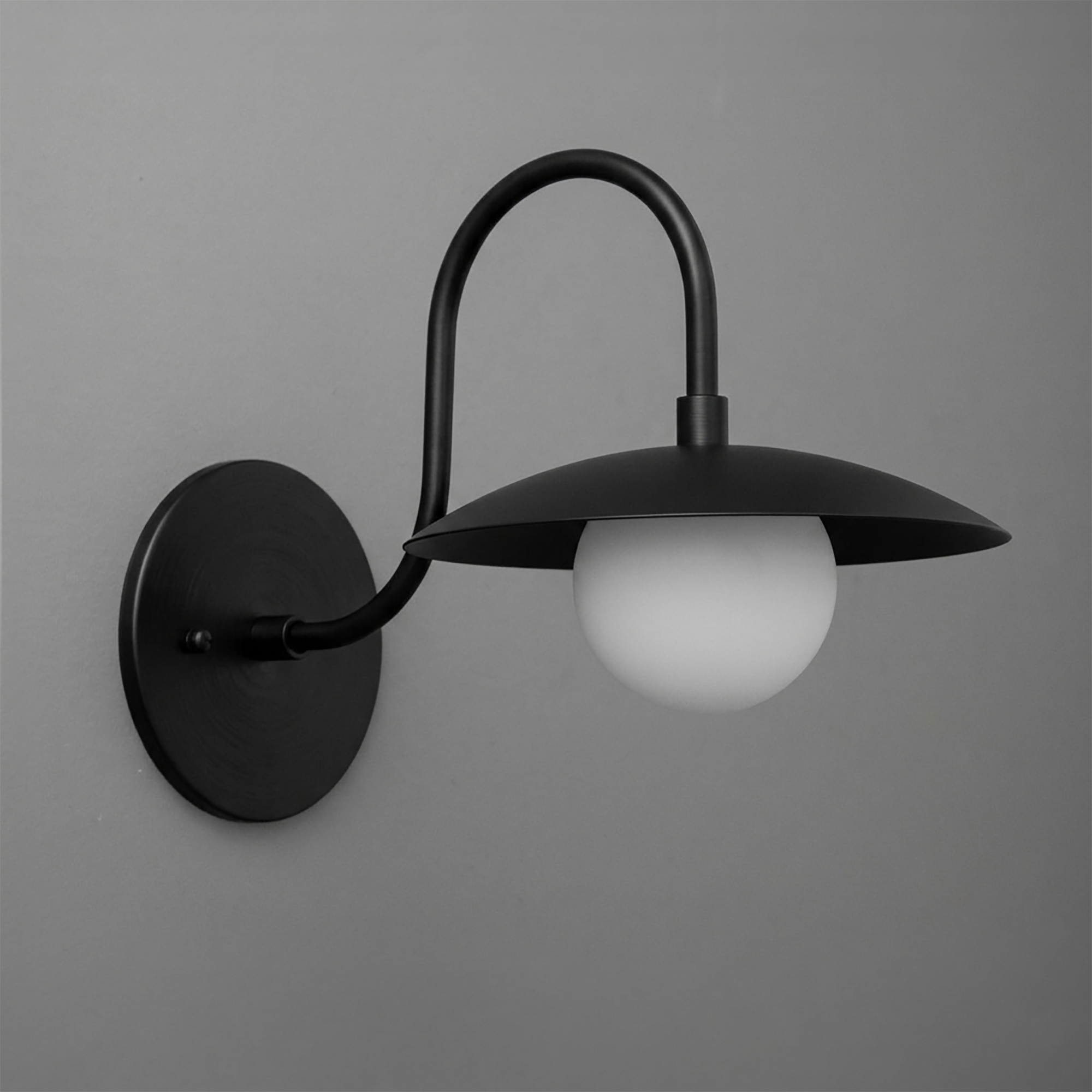 Black metal wall sconce with curved arm and wide flat shade, frosted globe bulb underneath. - SCONCE MODEL NO. 3799