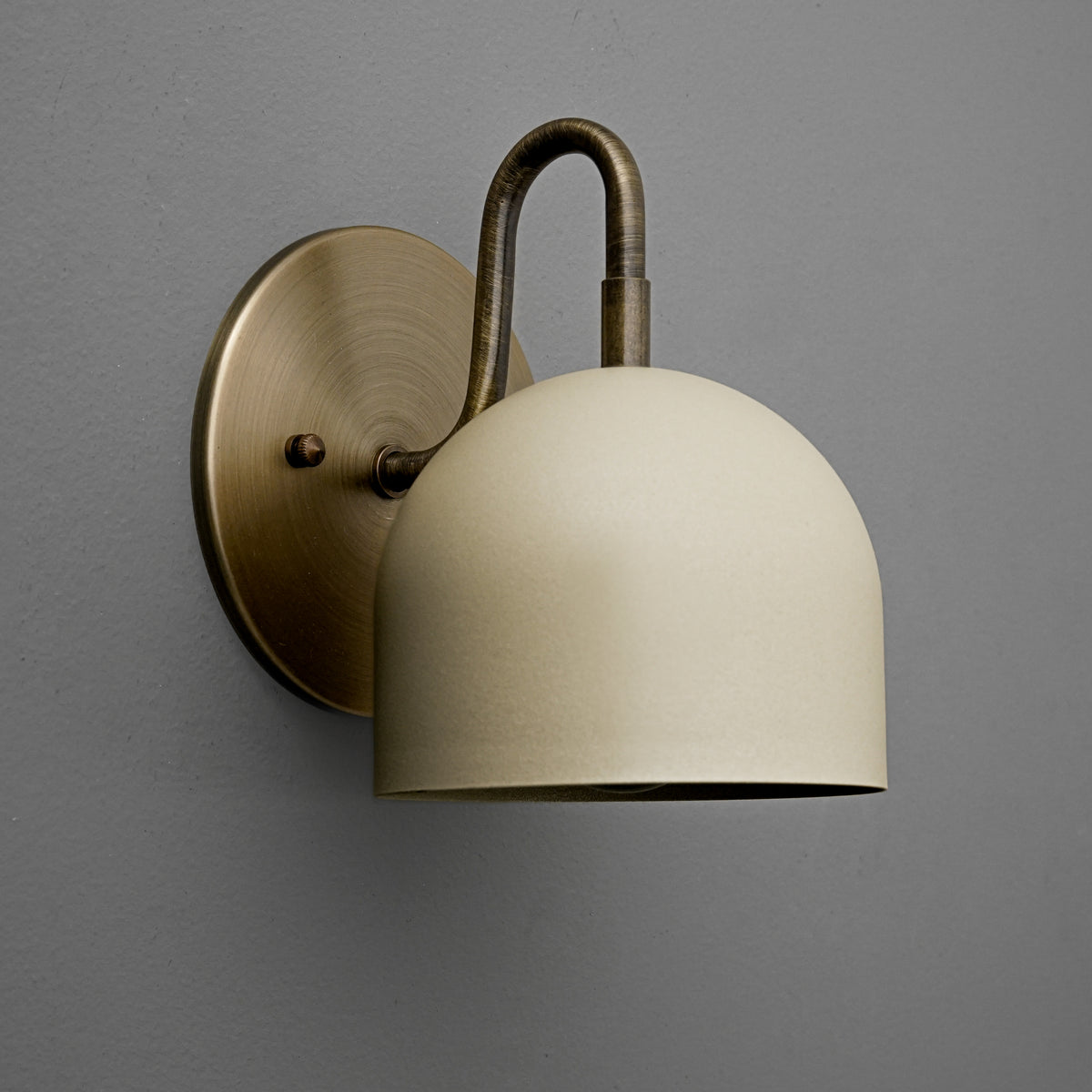 Antique brass wall sconce with smooth cream dome shade and curved arm on gray wall. - Sconce Model No. 5416