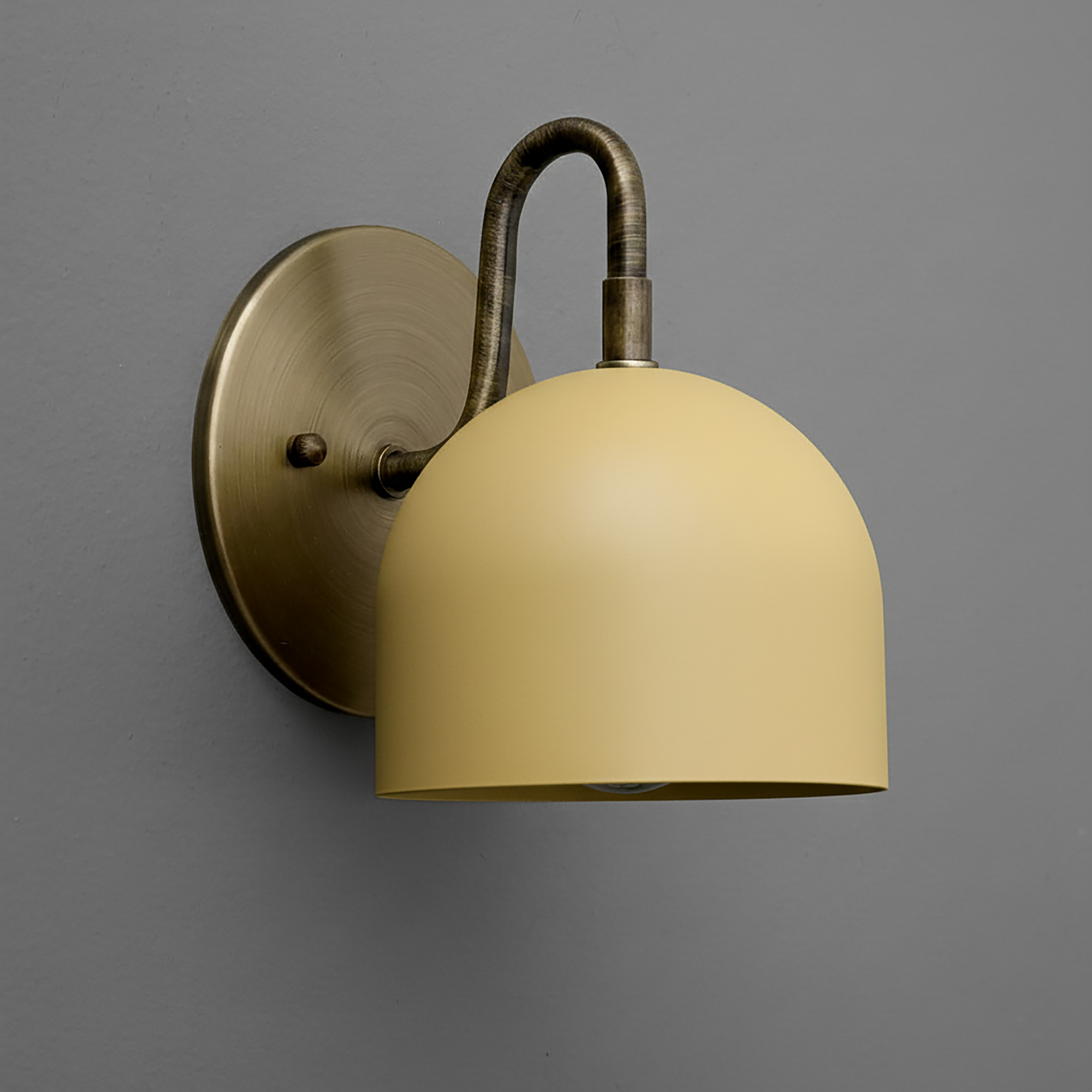 Matte mustard yellow dome-shaped metal sconce with antique brass curved arm and round backplate on gray wall. - Sconce Model No. 5416