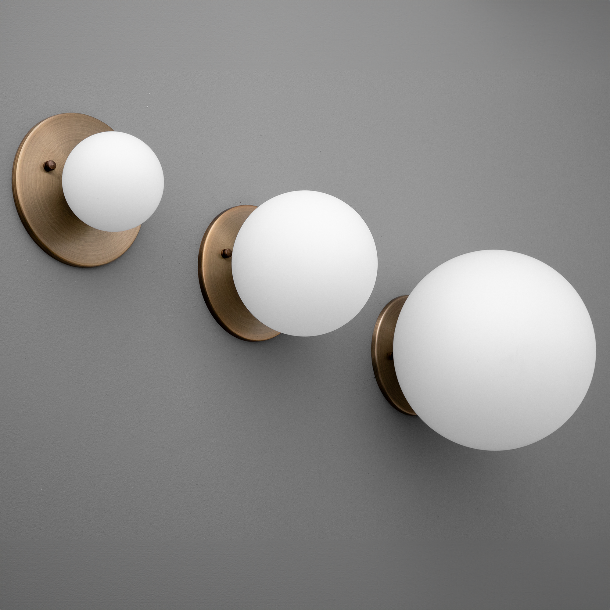 Set of three antique brass wall sconces with frosted white glass globes in varying sizes on gray background. - Sconce Model No. 5206