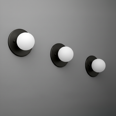 Set of three black metal sconces with white globe shades in small, medium, and large sizes mounted on a gray wall. - Sconce Model No. 5206