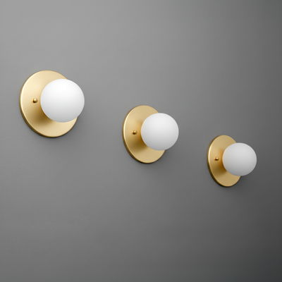 Three antique brass sconces with matte white globes mounted on a gray wall, varying sizes left to right. - Sconce Model No. 5206