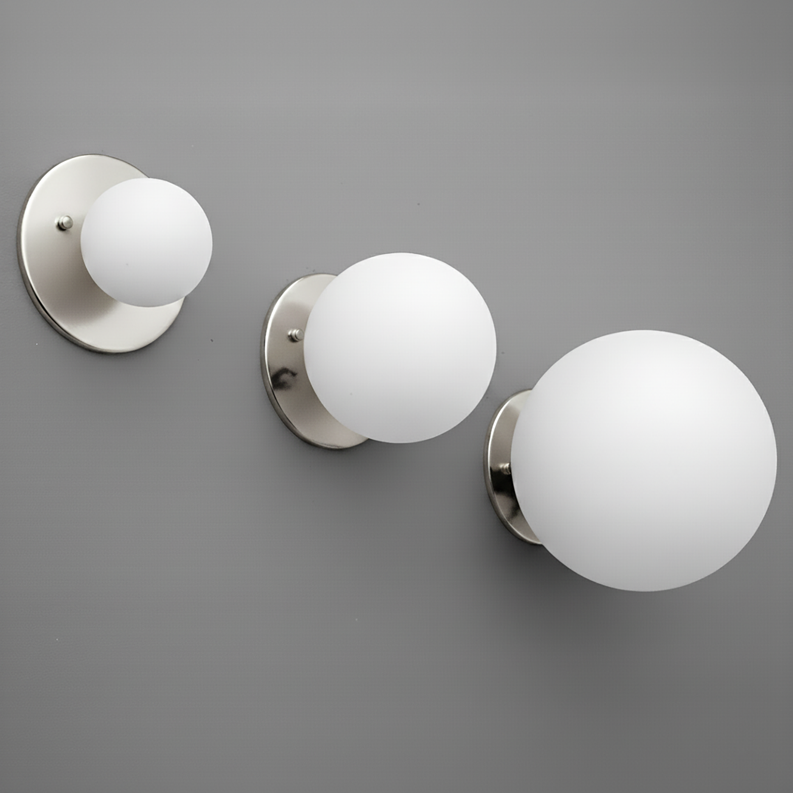 Three polished nickel wall sconces with round white glass globes in small, medium, and large sizes mounted on a gray wall. - Sconce Model No. 5206