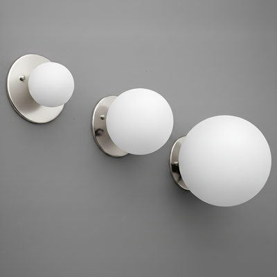 Three polished nickel wall sconces with round white glass globes in small, medium, and large sizes mounted on a gray wall. - Sconce Model No. 5206