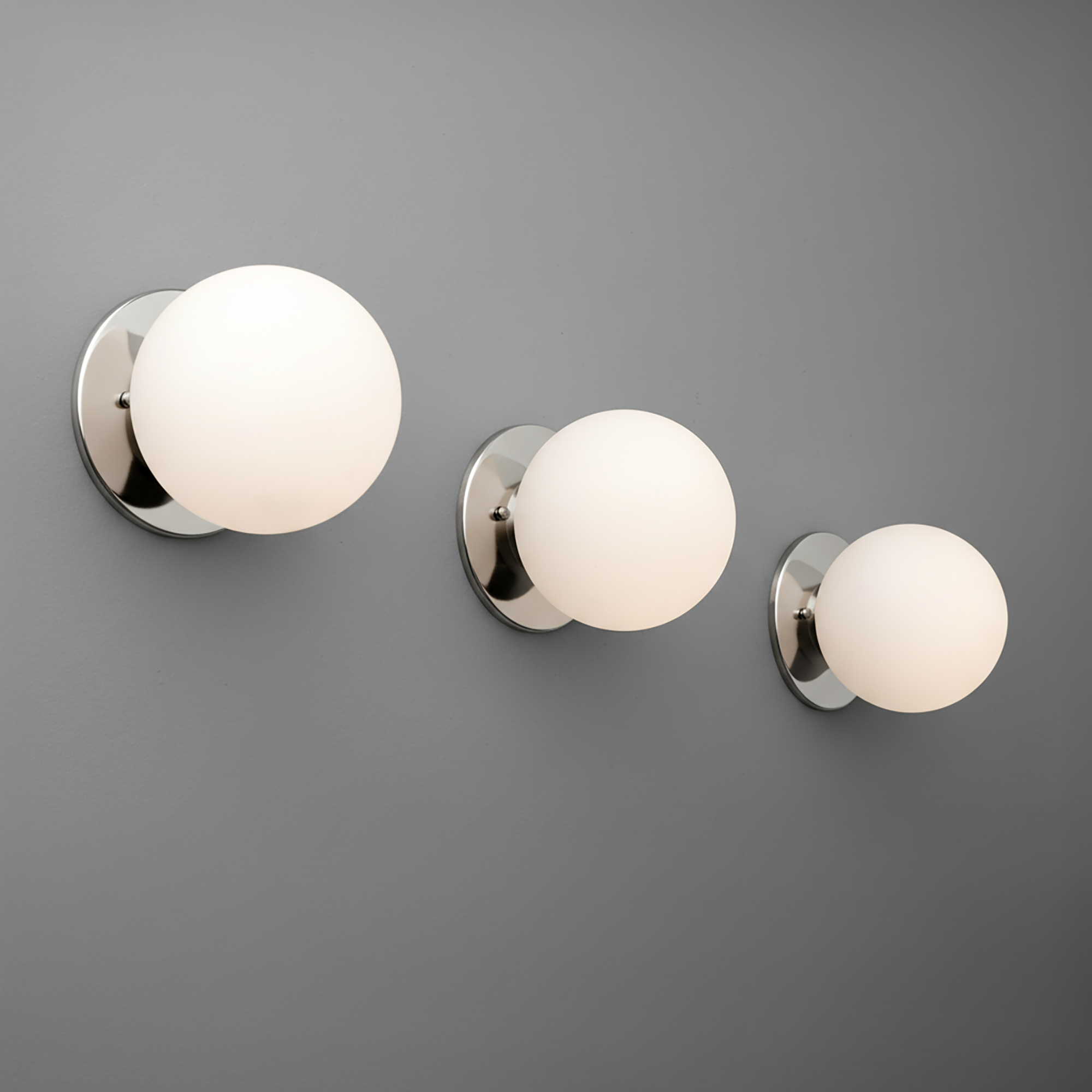 Set of three polished nickel wall sconces with frosted white glass globes in varying sizes mounted on gray wall. - Sconce Model No. 5206