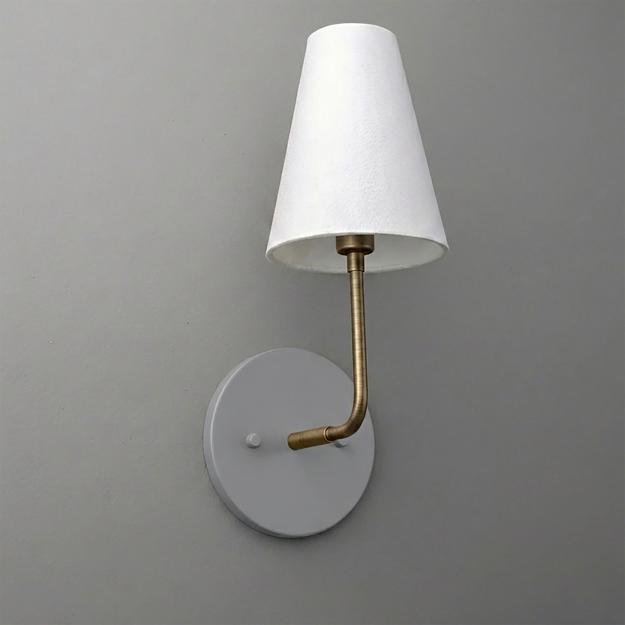 Wall-mounted sconce with round gray base, curved brass arm, and white tapered fabric shade. - SCONCE MODEL NO. 3928