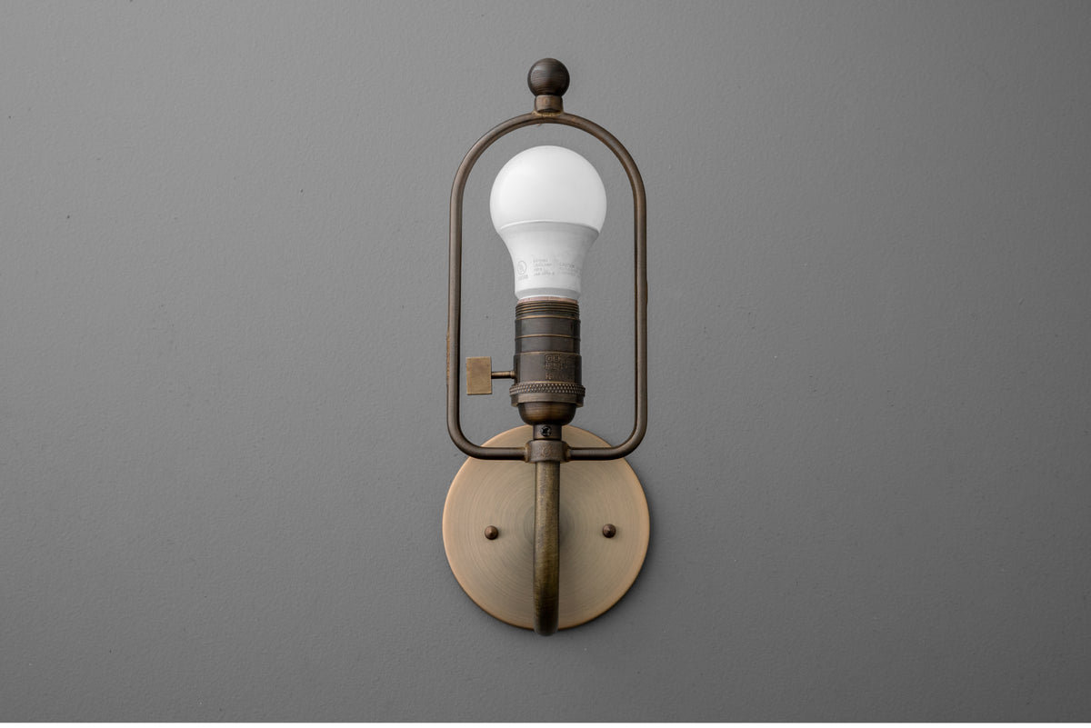Antique brass wall sconce with exposed bulb and rounded rectangular frame on gray background. - SCONCE MODEL NO. 0303