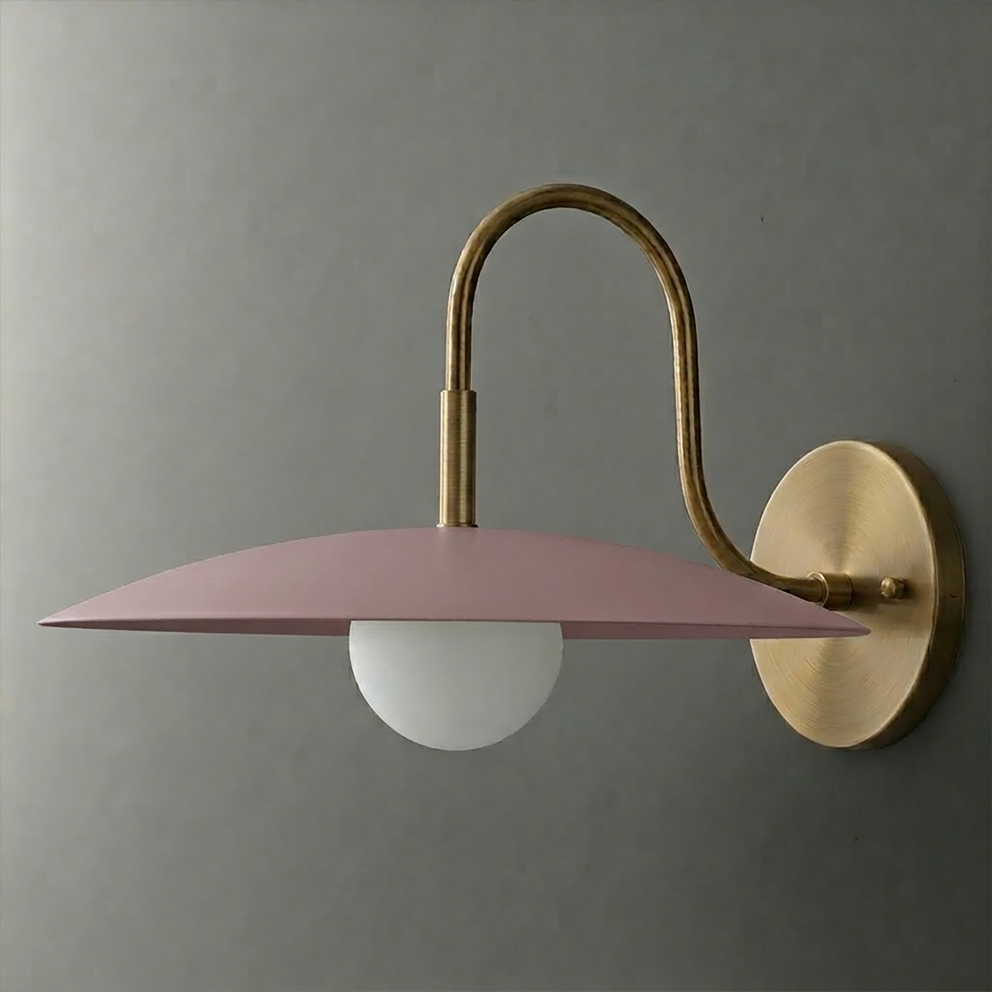 Wall sconce with brass curved arm and round base, featuring a flat matte pink metal shade and a white globe bulb underneath. - SCONCE MODEL NO. 9171