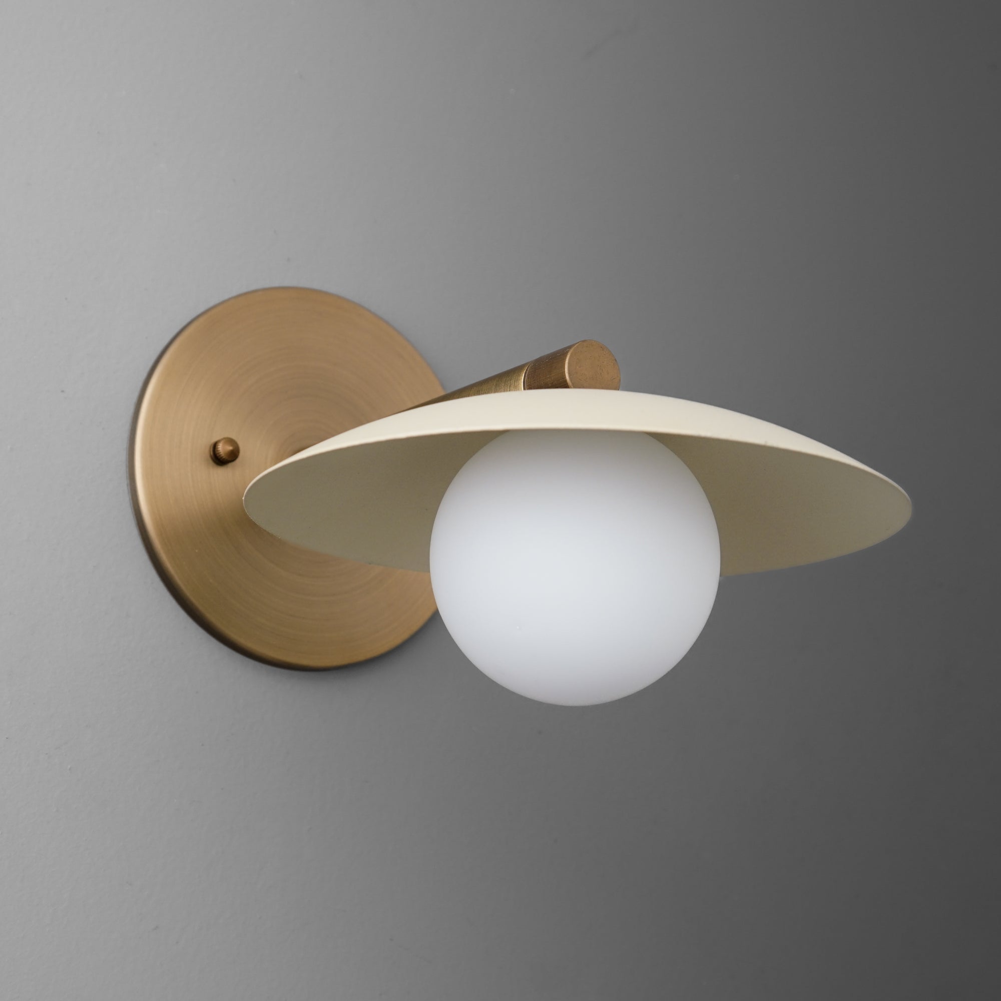 Wall sconce with matte brass circular base and arm, featuring a white opaque glass globe light under a flat cream metal. - SCONCE MODEL NO. 9872