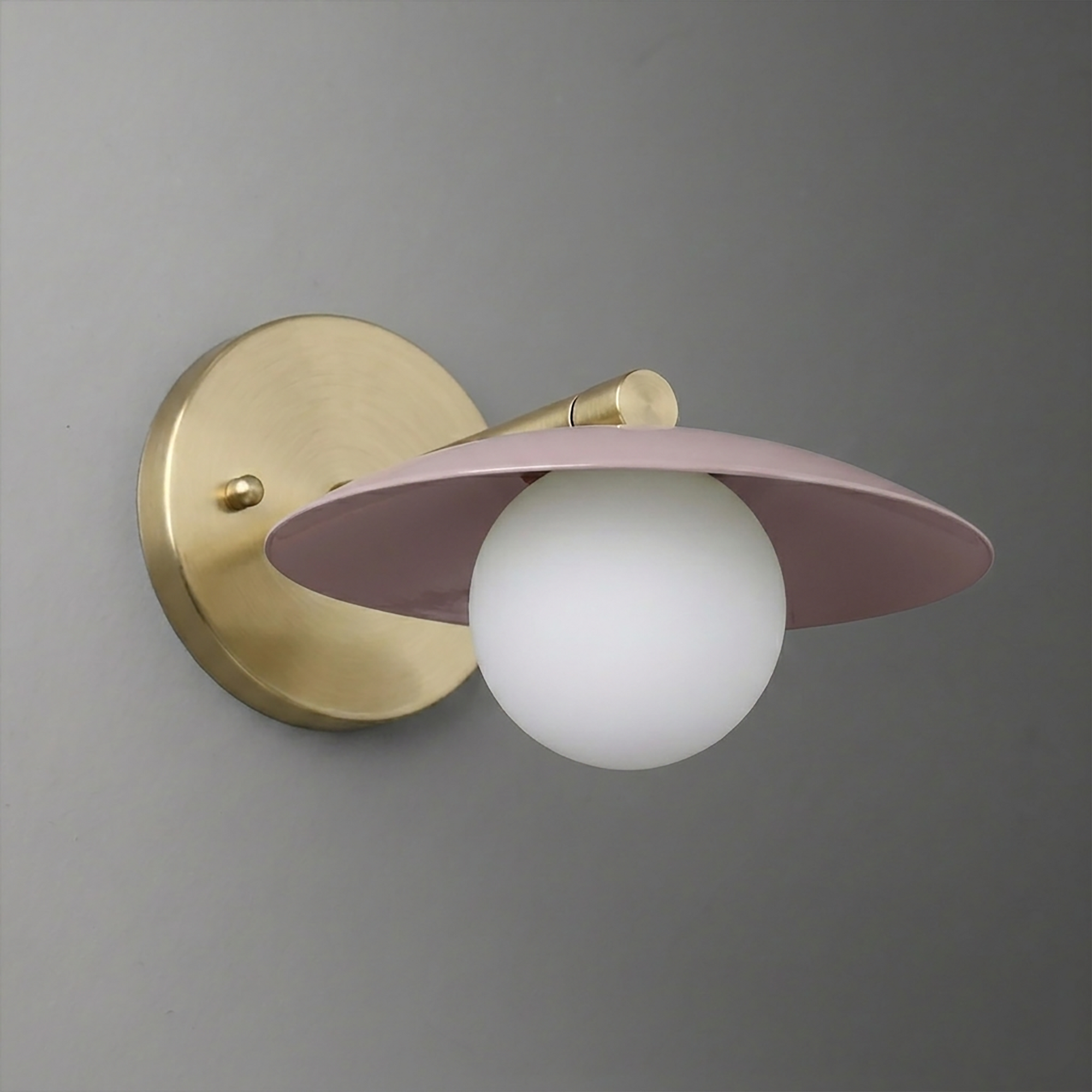 Wall sconce with matte gold round base and arm, featuring a frosted globe bulb under a flat dusty pink metal shade. - SCONCE MODEL NO. 9872