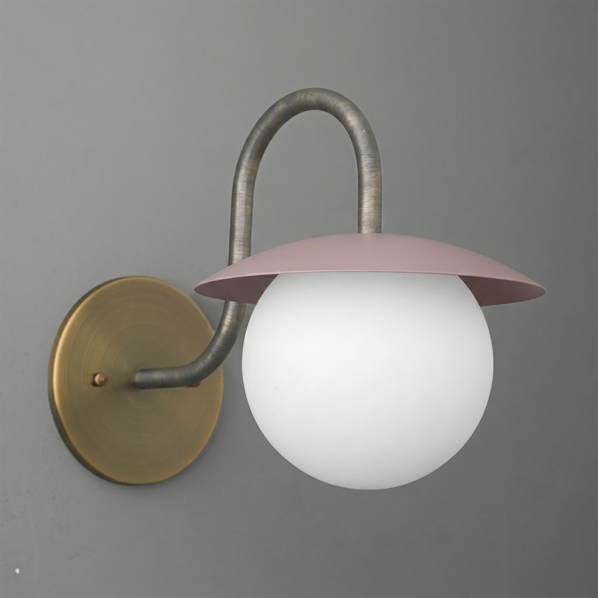 Wall-mounted sconce with a round brass base, curved metal arm, pink flat dome shade, and white globe bulb. - SCONCE MODEL NO. 2206