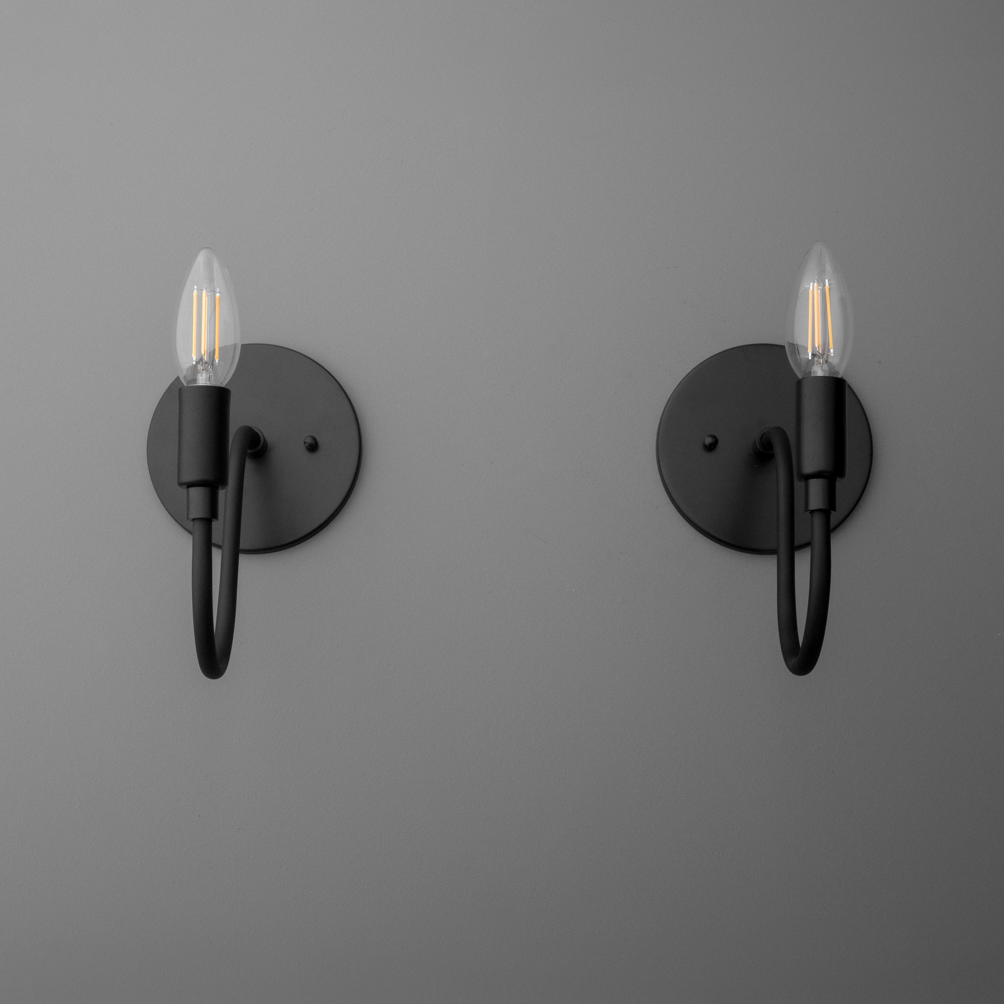 Pair of matte black metal wall sconces with minimalist curved arms and exposed candle-style bulbs. - SCONCE MODEL NO. 7321