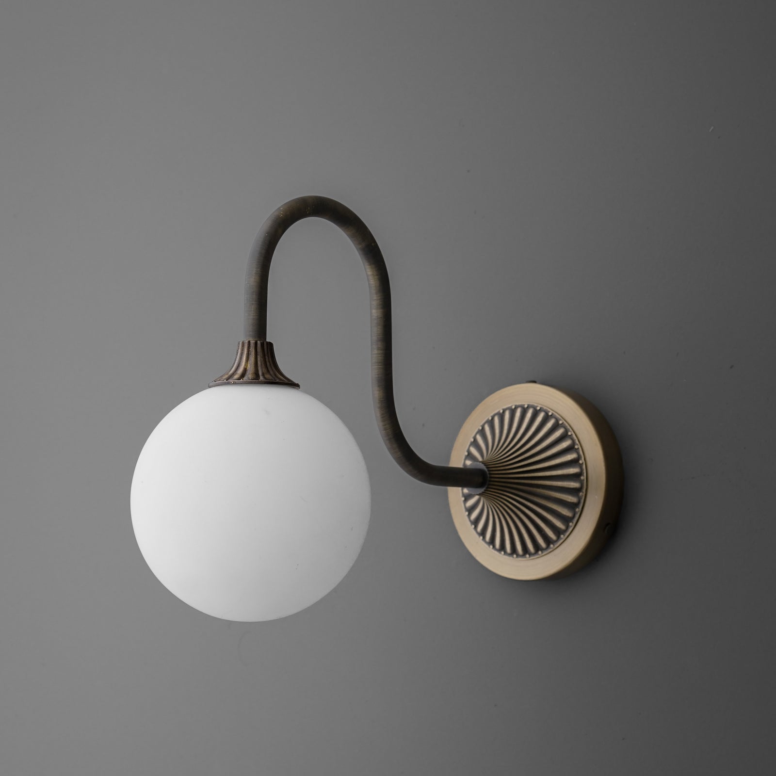 Wall sconce with a curved dark bronze arm and round white glass globe shade attached to a circular brass backplate. - SCONCE MODEL NO. 1085