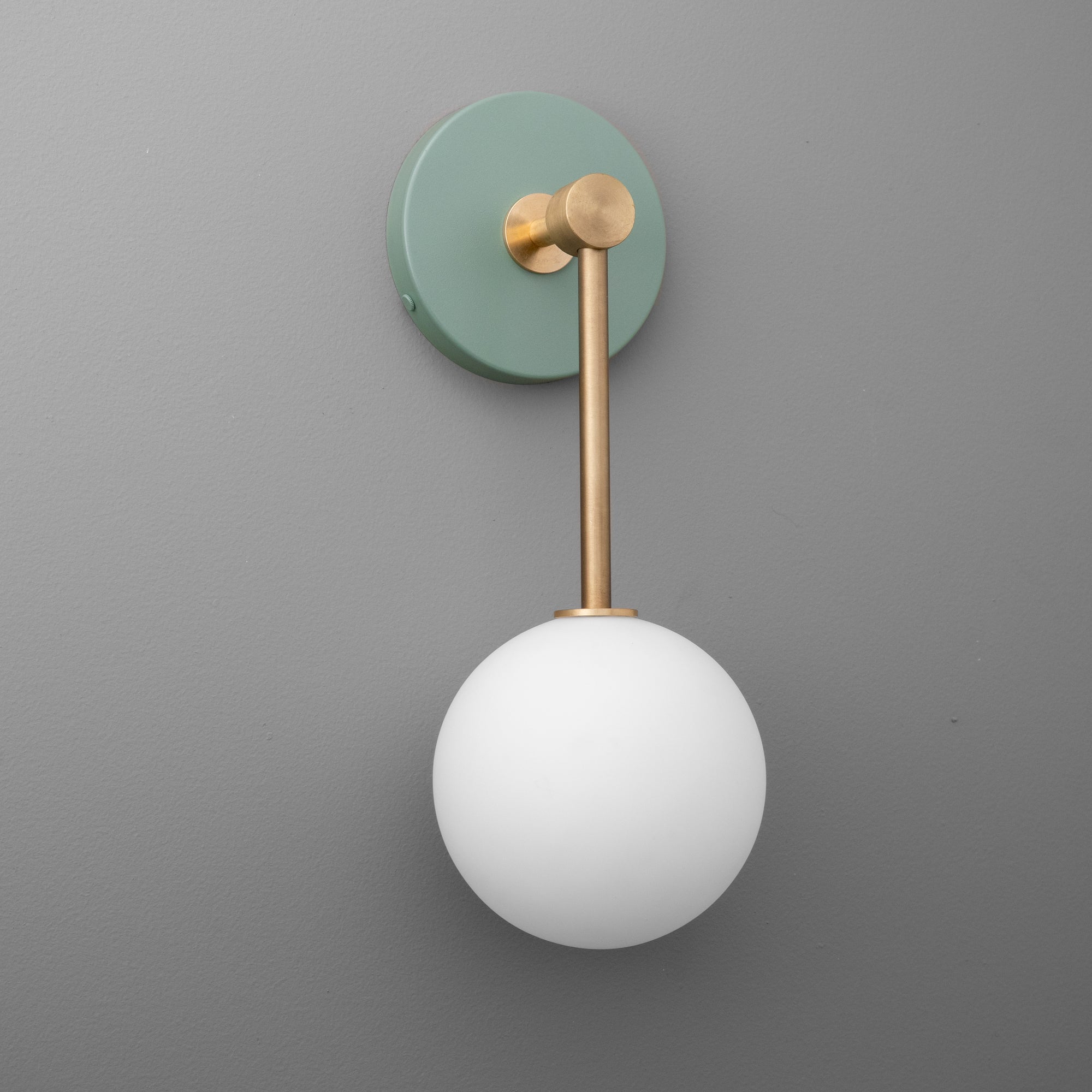 Wall sconce with white globe shade and raw brass cylindrical arm on mint green round mounting plate. - Sconce Model No. 5999