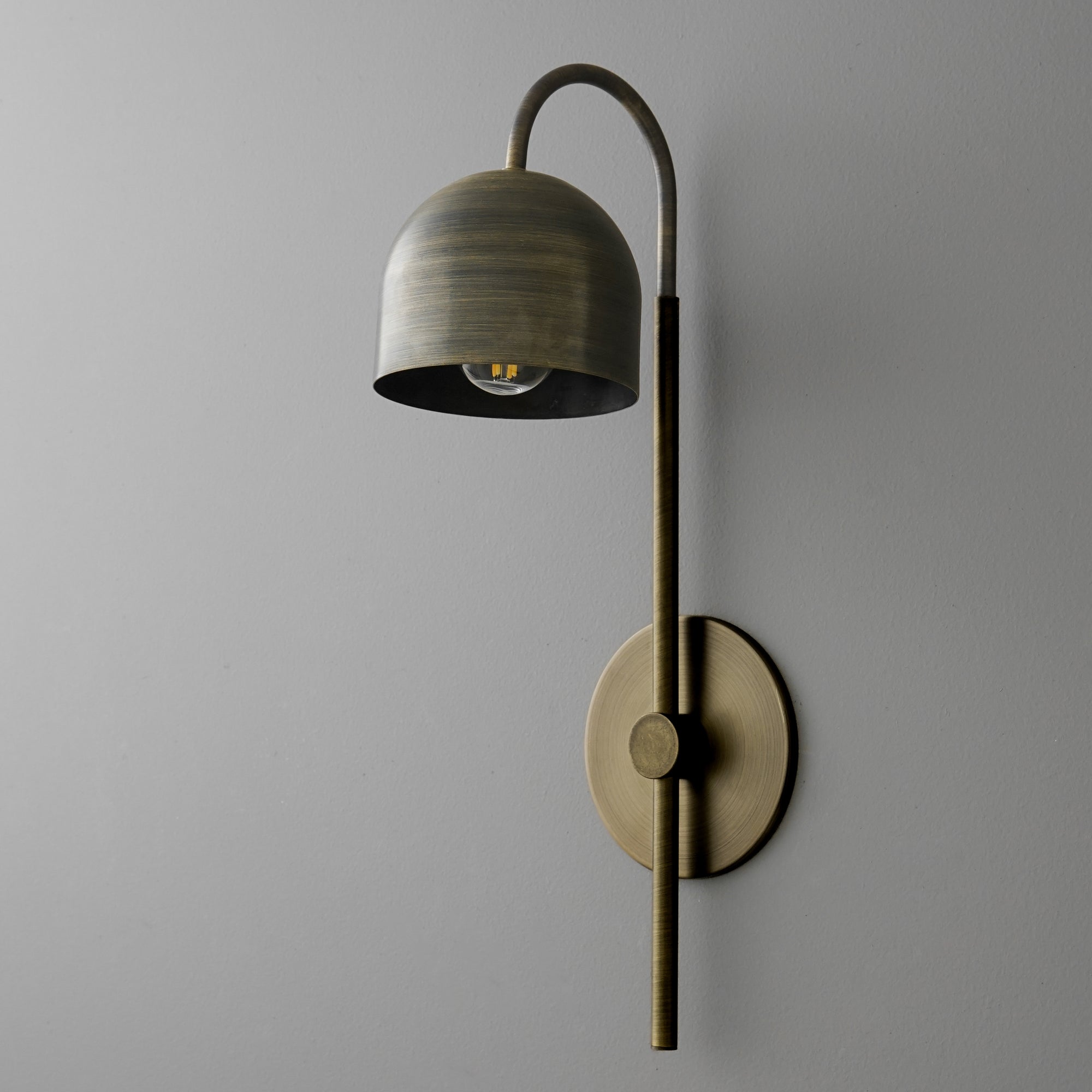 Antique brass wall sconce with curved arm and dome-shaped shade against gray wall. - Sconce Model No. 7340