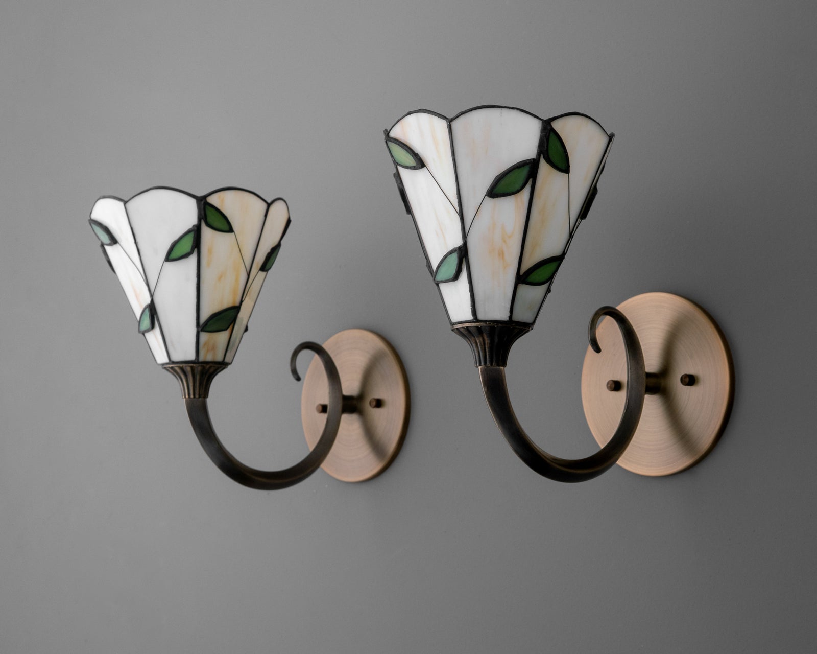Pair of wall sconces with curved black metal arms and stained glass shades featuring white panels and green leaf accents. - SCONCE MODEL NO. 1056