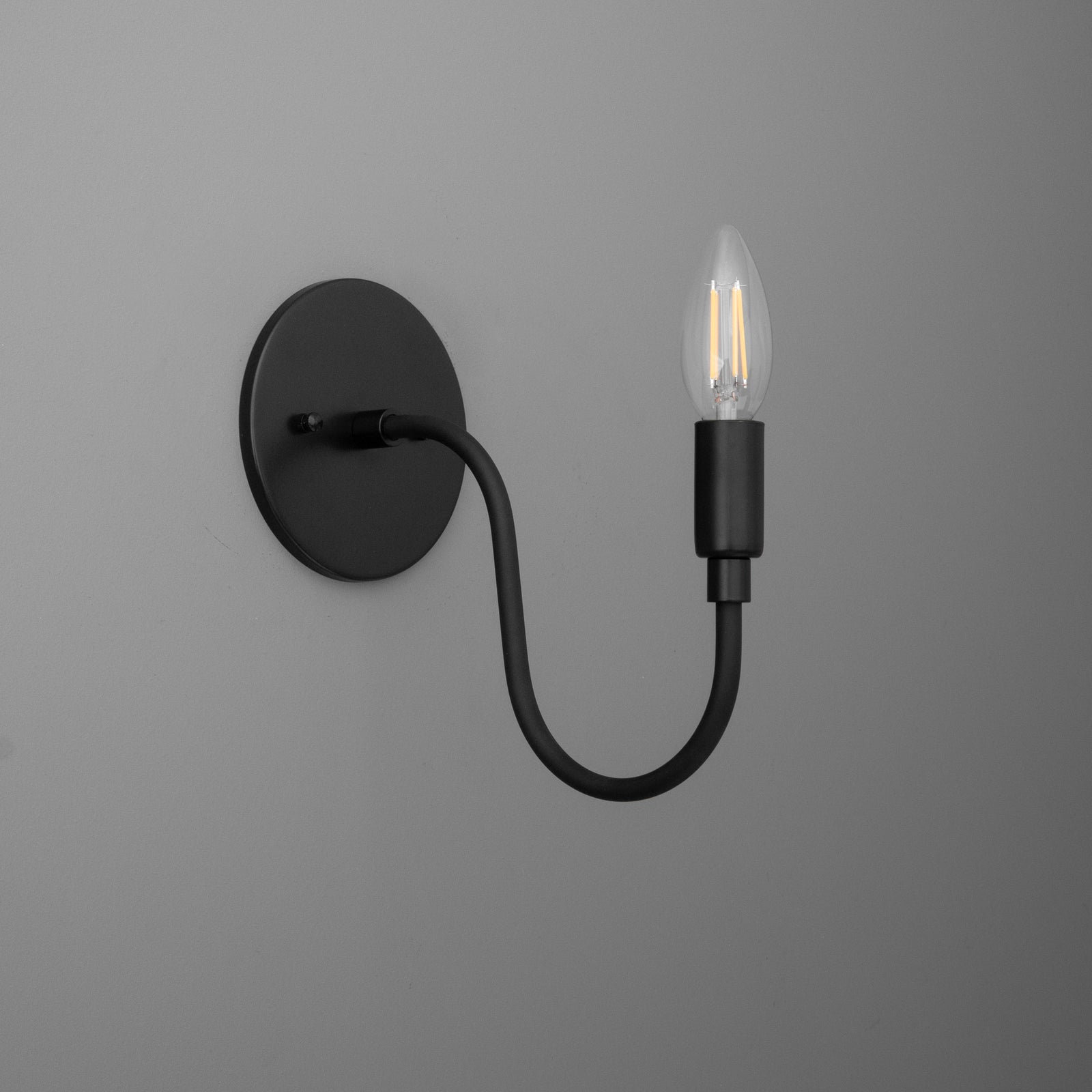 Matte black metal wall sconce with round base and curved arm holding a single exposed vintage-style bulb. - SCONCE MODEL NO. 7321