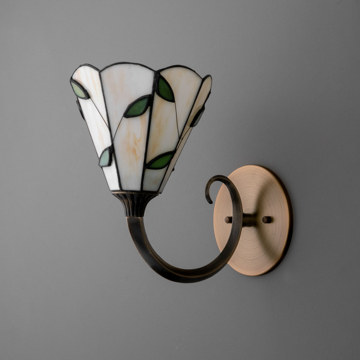 Wall sconce with brass round base and curved black metal arm holding a stained glass shade with white panels and green leaf. - SCONCE MODEL NO. 1056