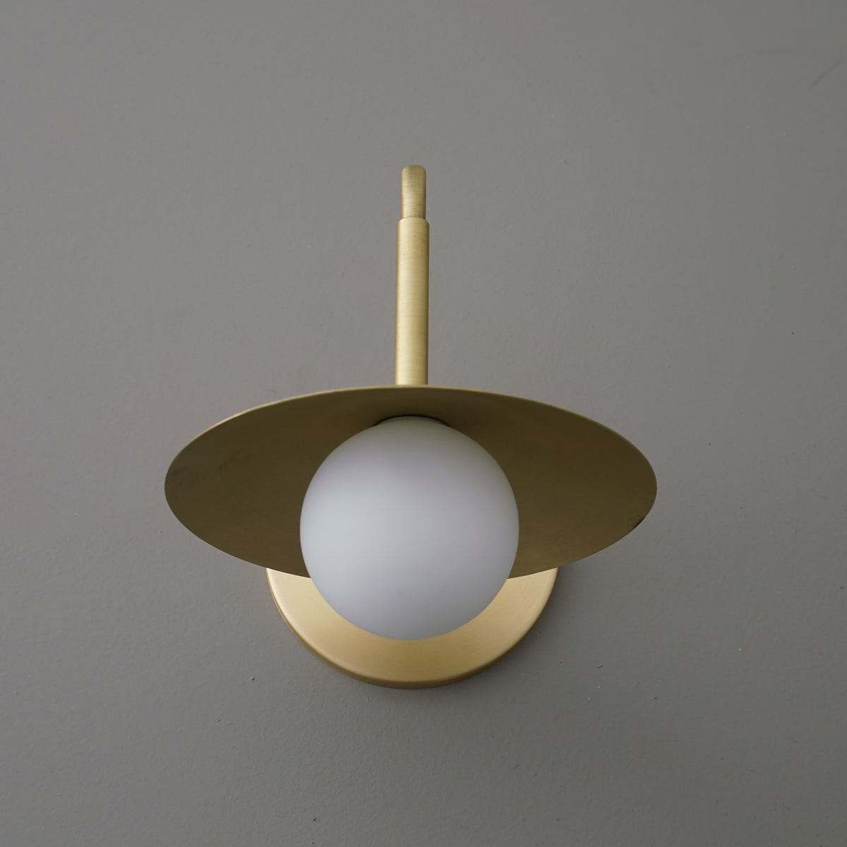 Matte gold metal sconce with flat circular shade and frosted white globe bulb centered beneath. - SCONCE MODEL NO. 1013