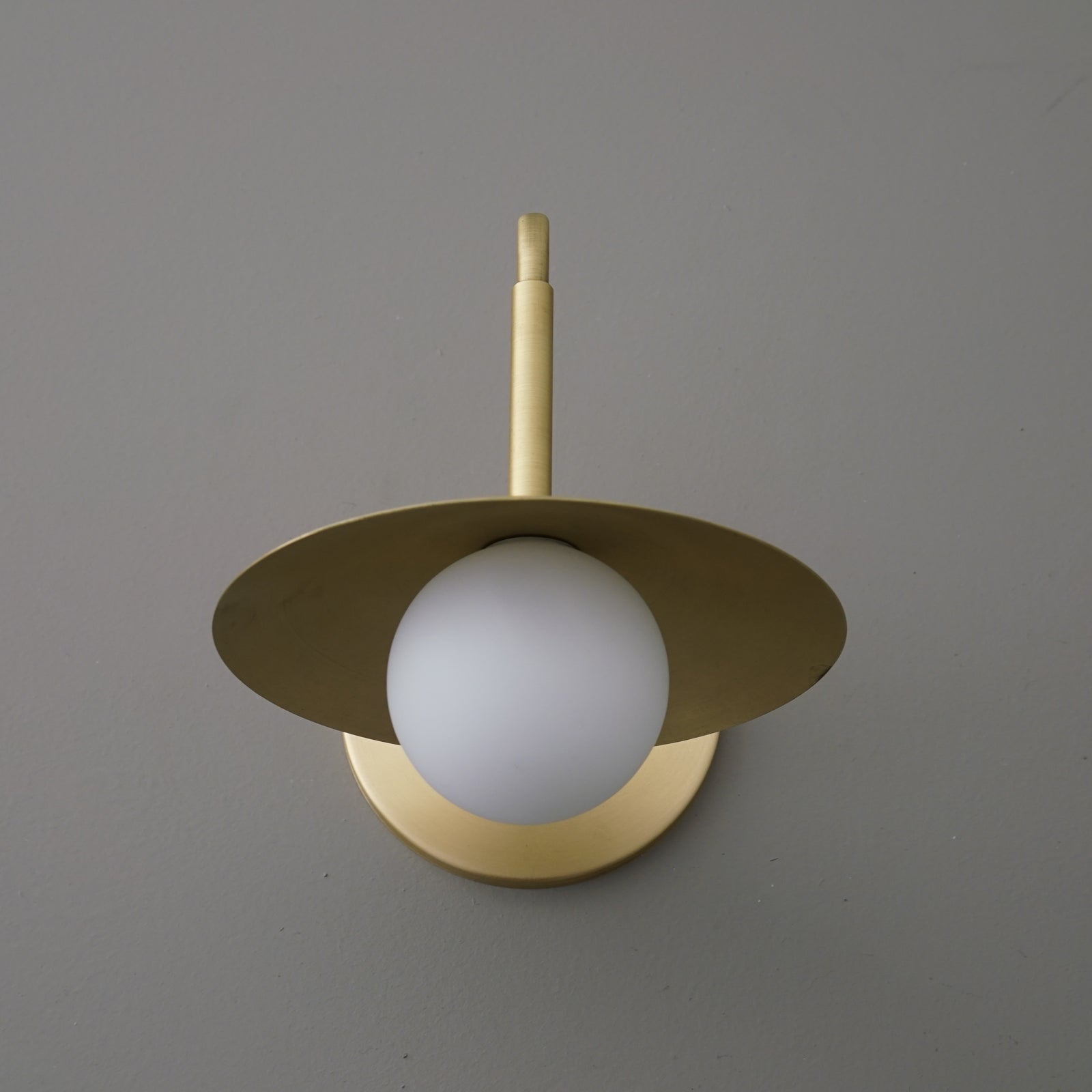 Matte gold metal sconce with flat circular shade and frosted white globe bulb centered beneath. - SCONCE MODEL NO. 1013