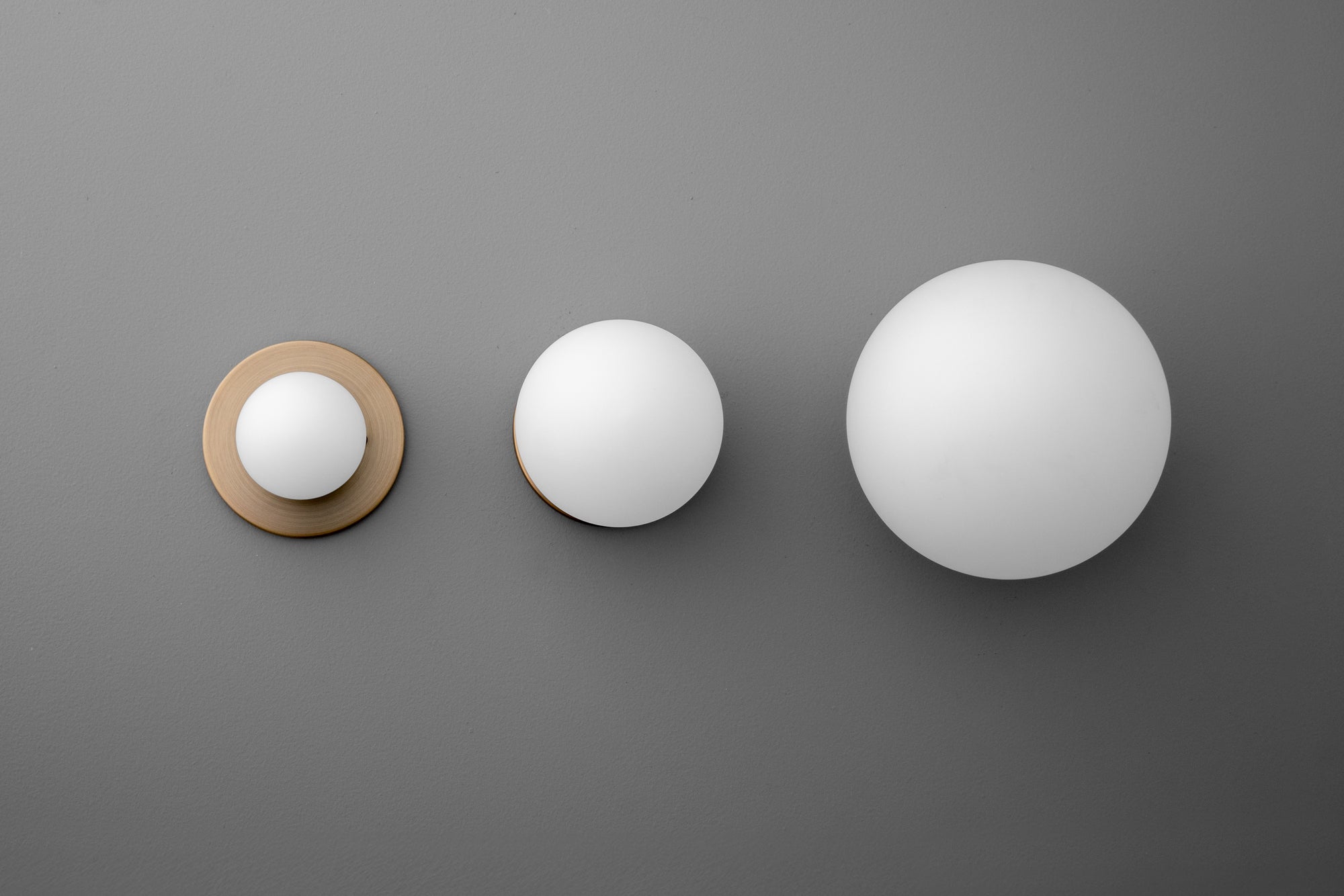Three brass wall sconces with white globe shades in small, medium, and large sizes mounted on a gray wall. - Sconce Model No. 5206