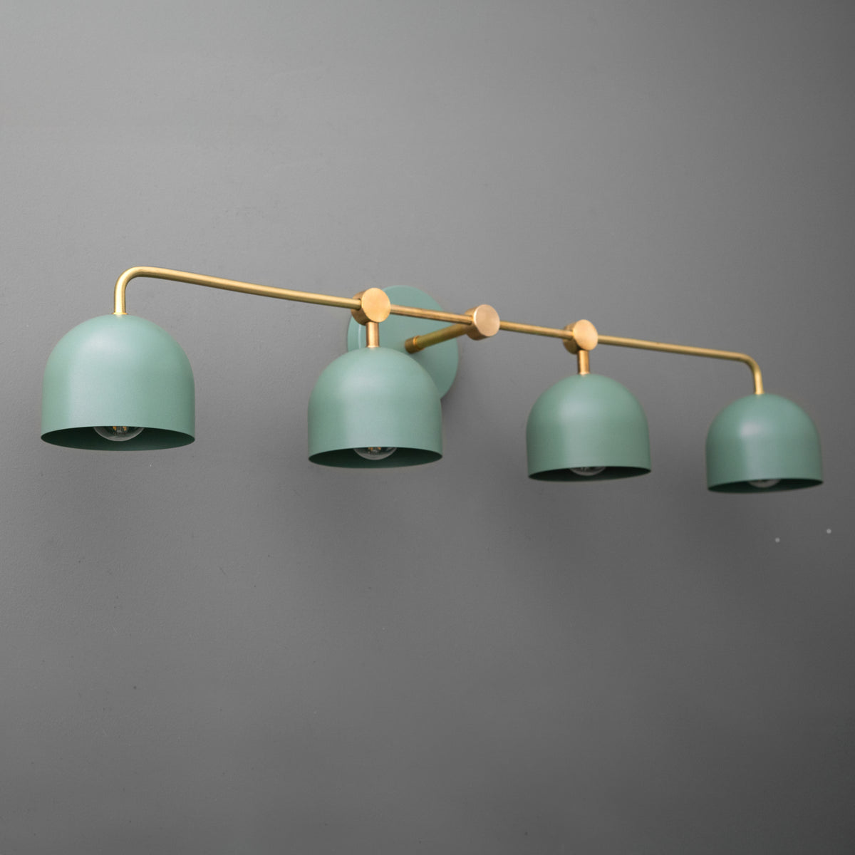 Angled view of a four-light vanity light in Raw or Sealed Brass with Green dome shades hanging from a long bar. - VANITY LIGHT MODEL NO. 1124