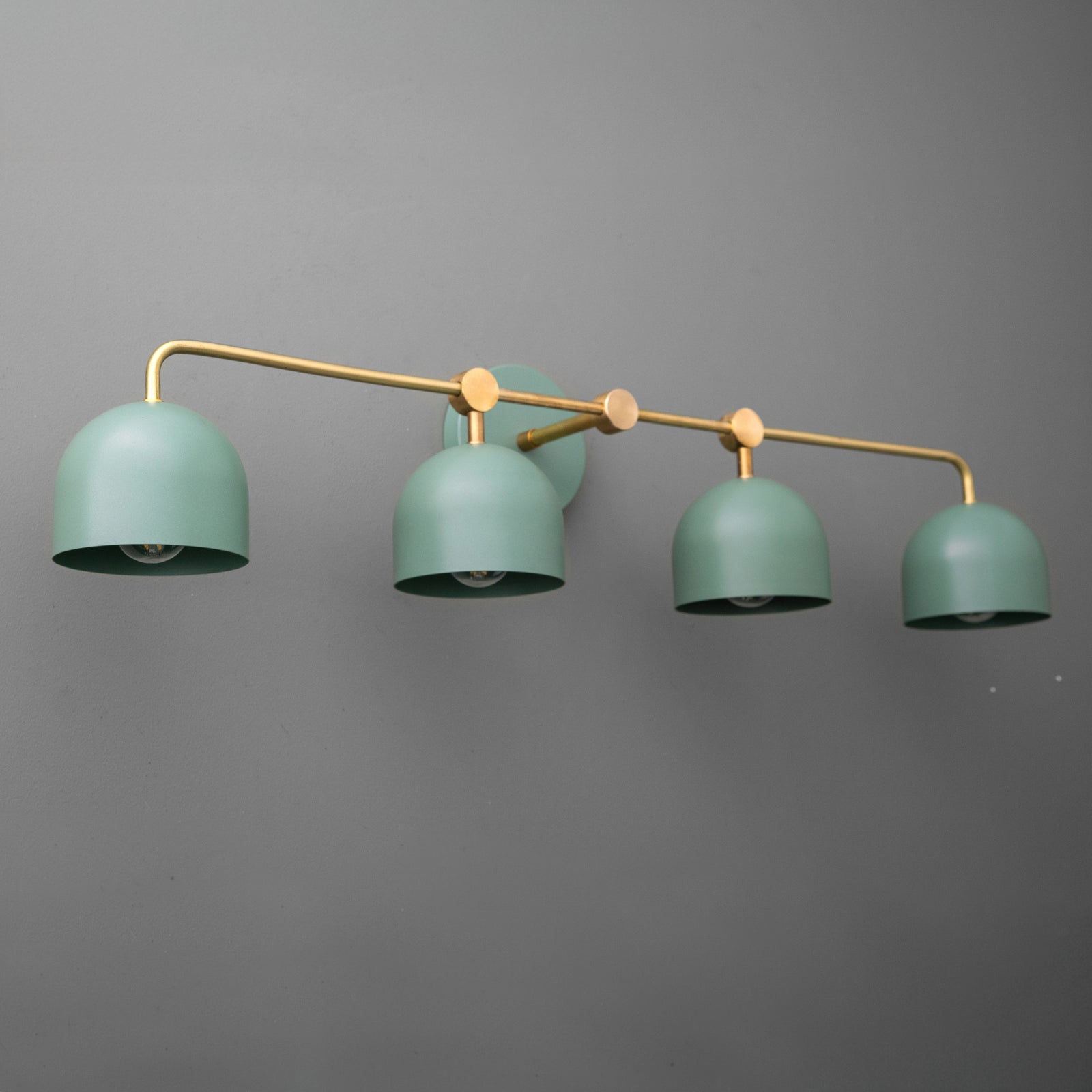 Angled view of a four-light vanity light in Raw or Sealed Brass with Green dome shades hanging from a long bar. - VANITY LIGHT MODEL NO. 1124