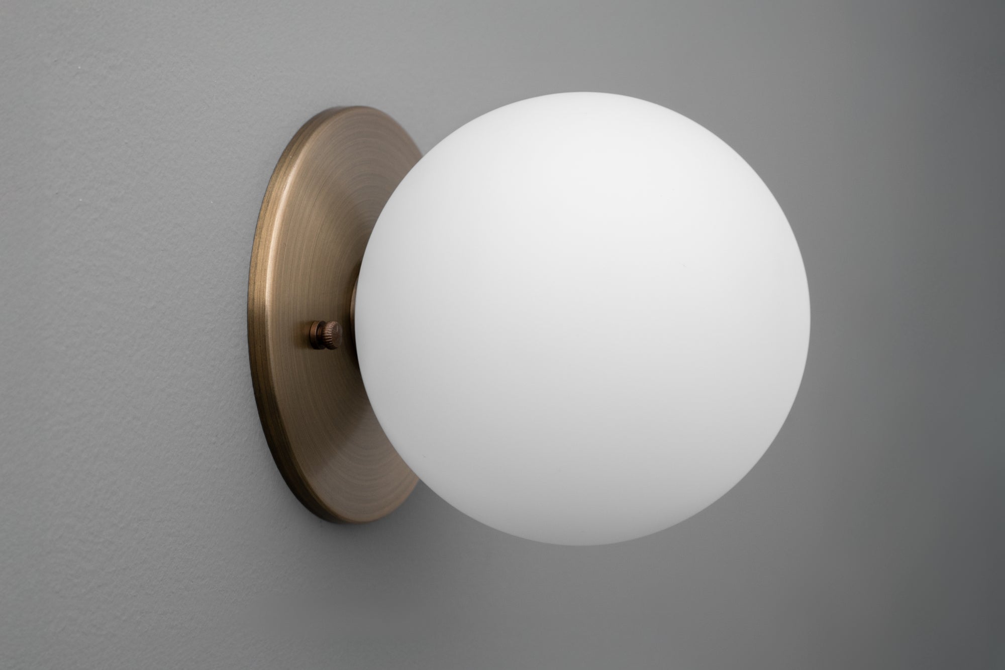 Antique brass wall sconce with frosted white 5-inch globe shade on gray wall. - Sconce Model No. 5206