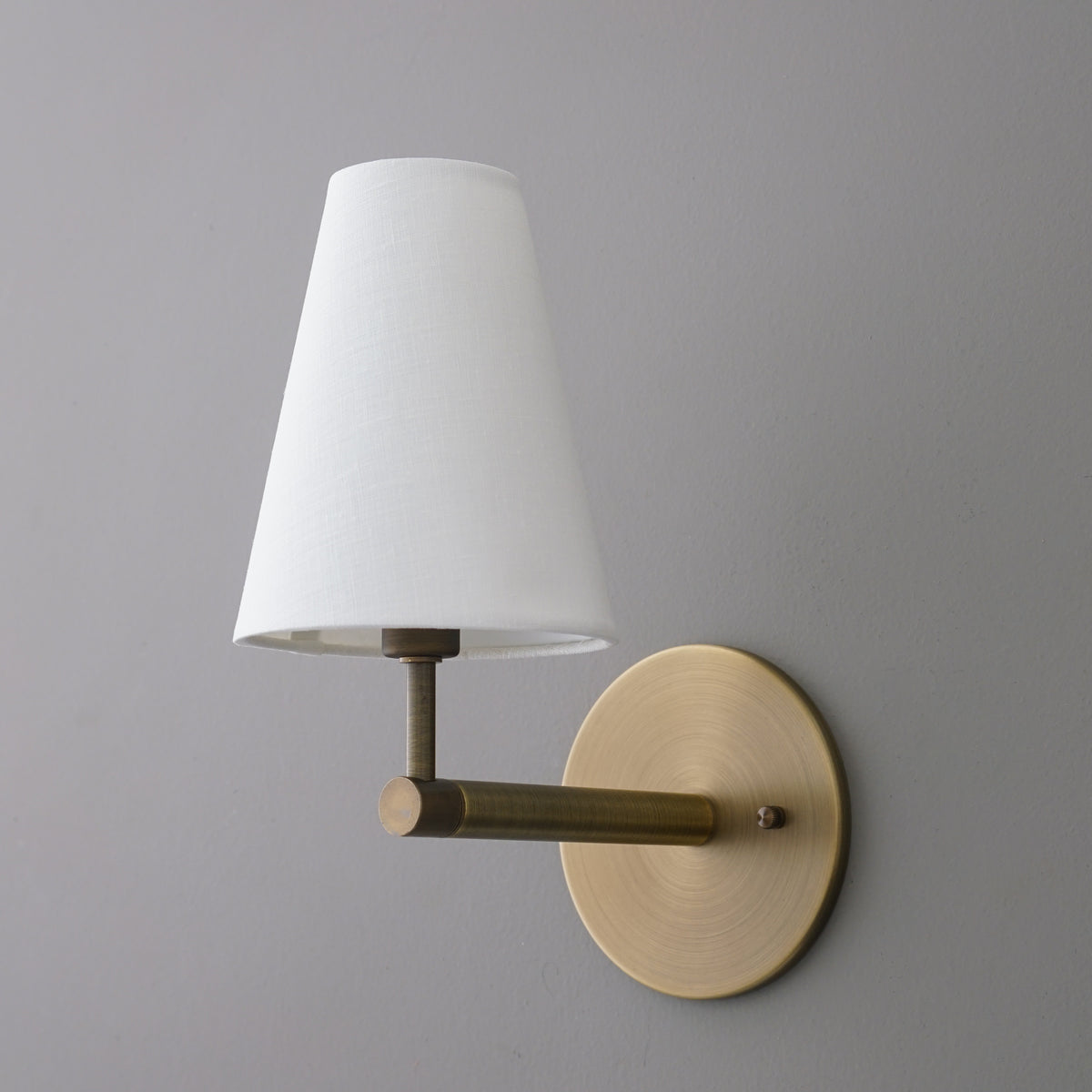 Wall-mounted sconce with a round brass base and arm, featuring a white tapered fabric shade. - SCONCE MODEL NO. 1014