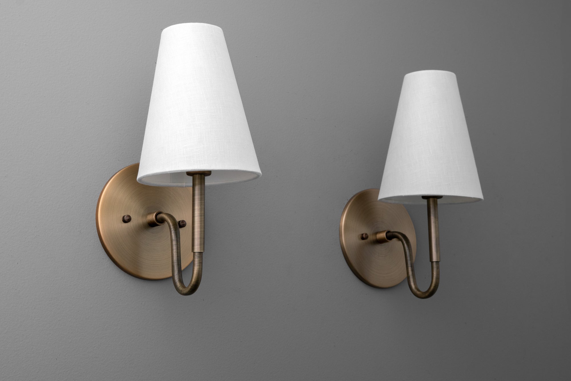 Pair of wall sconces with white tapered fabric shades and antique brass curved arms mounted on round backplates. - SCONCE MODEL NO. 1699