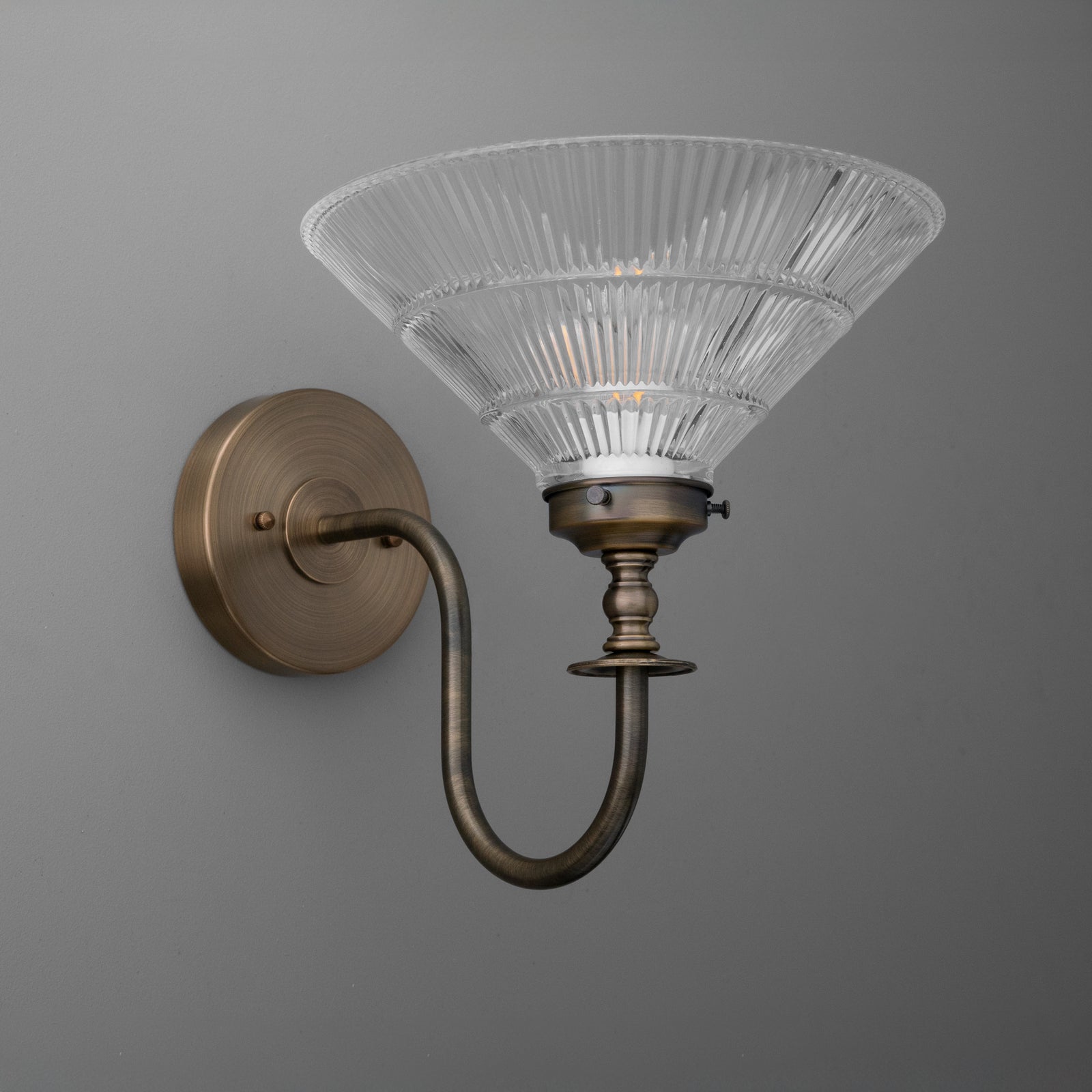 Antique brass sconce with curved arm and ribbed clear glass shade in flared cone shape. - SCONCE MODEL NO. 8036