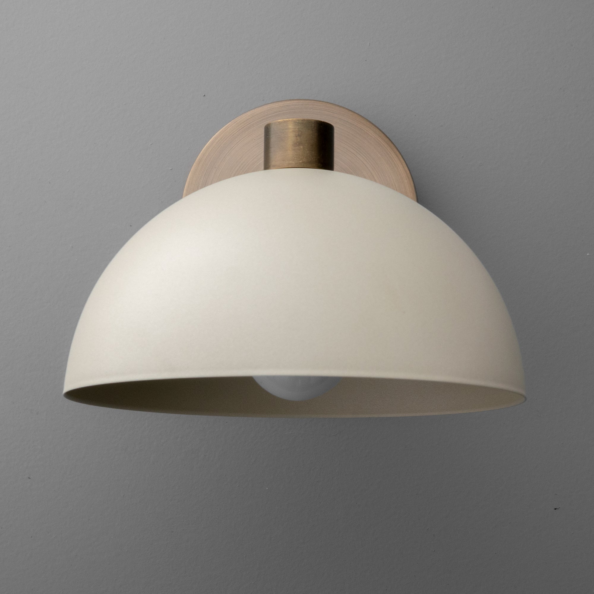 Cream dome-shaped sconce with antique brass circular wall mount and visible light bulb. - SCONCE MODEL NO. 2946