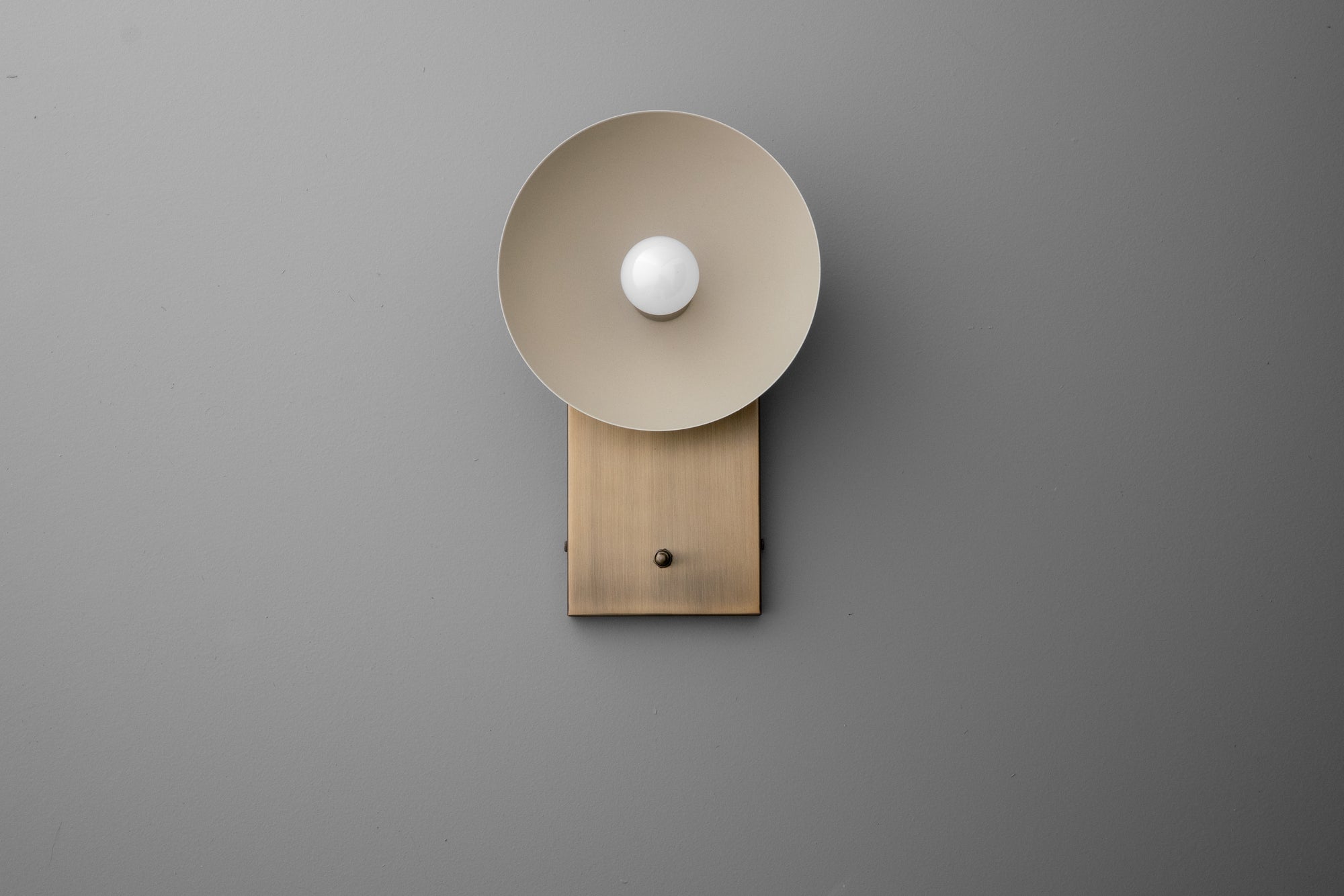 Wall sconce with cream dome shade and raw brass rectangular backplate on gray wall. - SCONCE MODEL NO. 3357