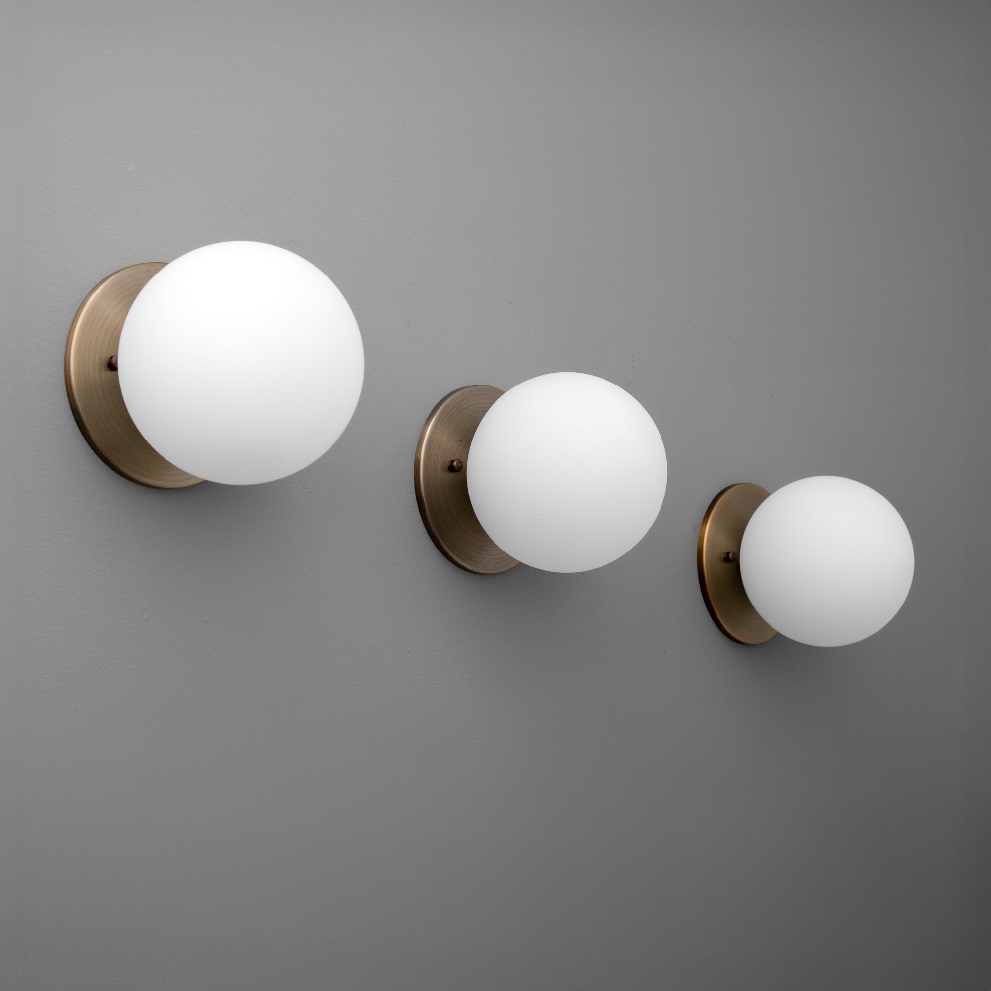 Three antique brass wall sconces with white frosted glass globes in small, medium, and large sizes on a gray wall. - Sconce Model No. 5206